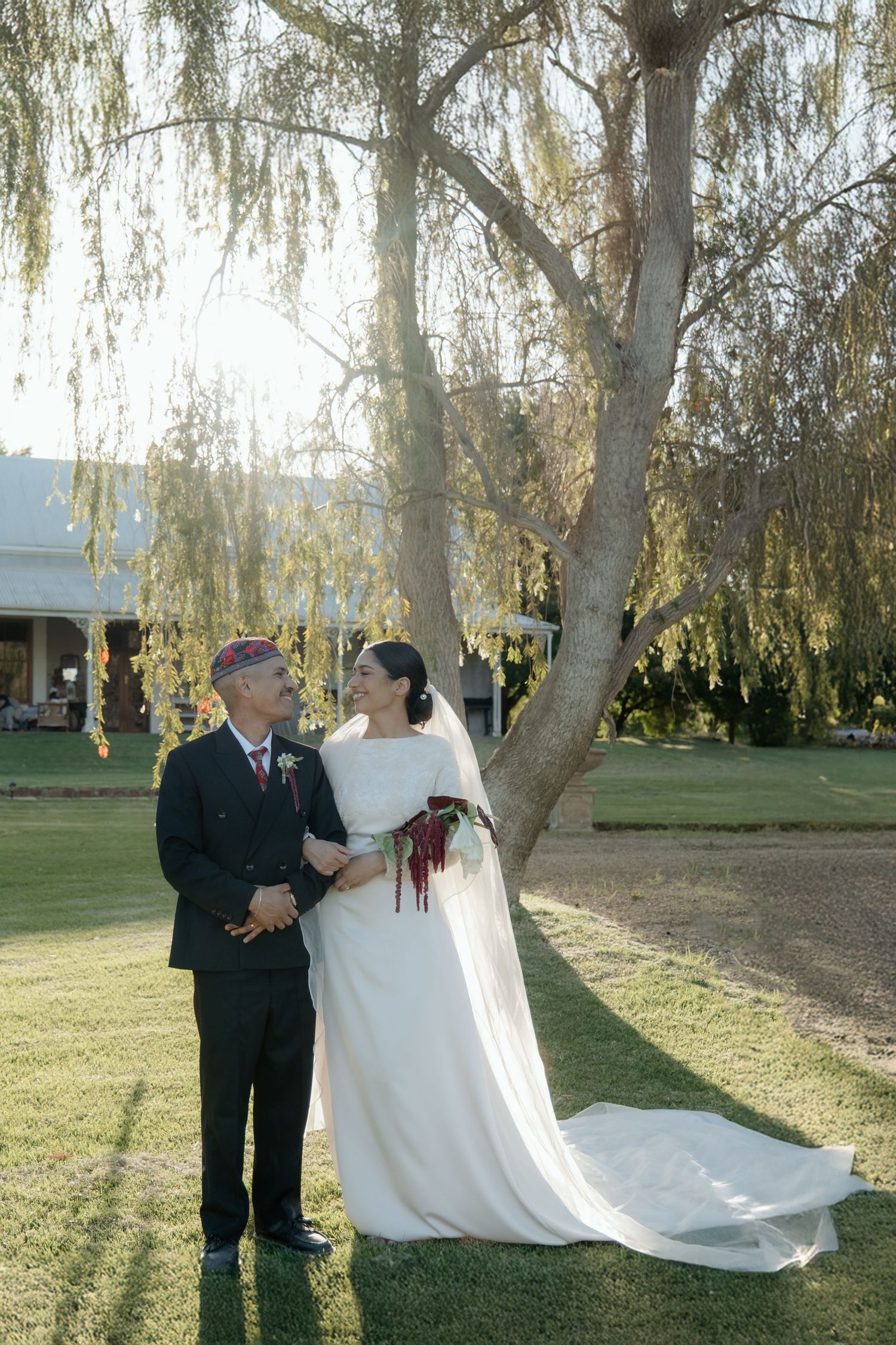 Newlywed couple stands beneath a weeping willow tree on a sunny day at an outdoor wedding venue.