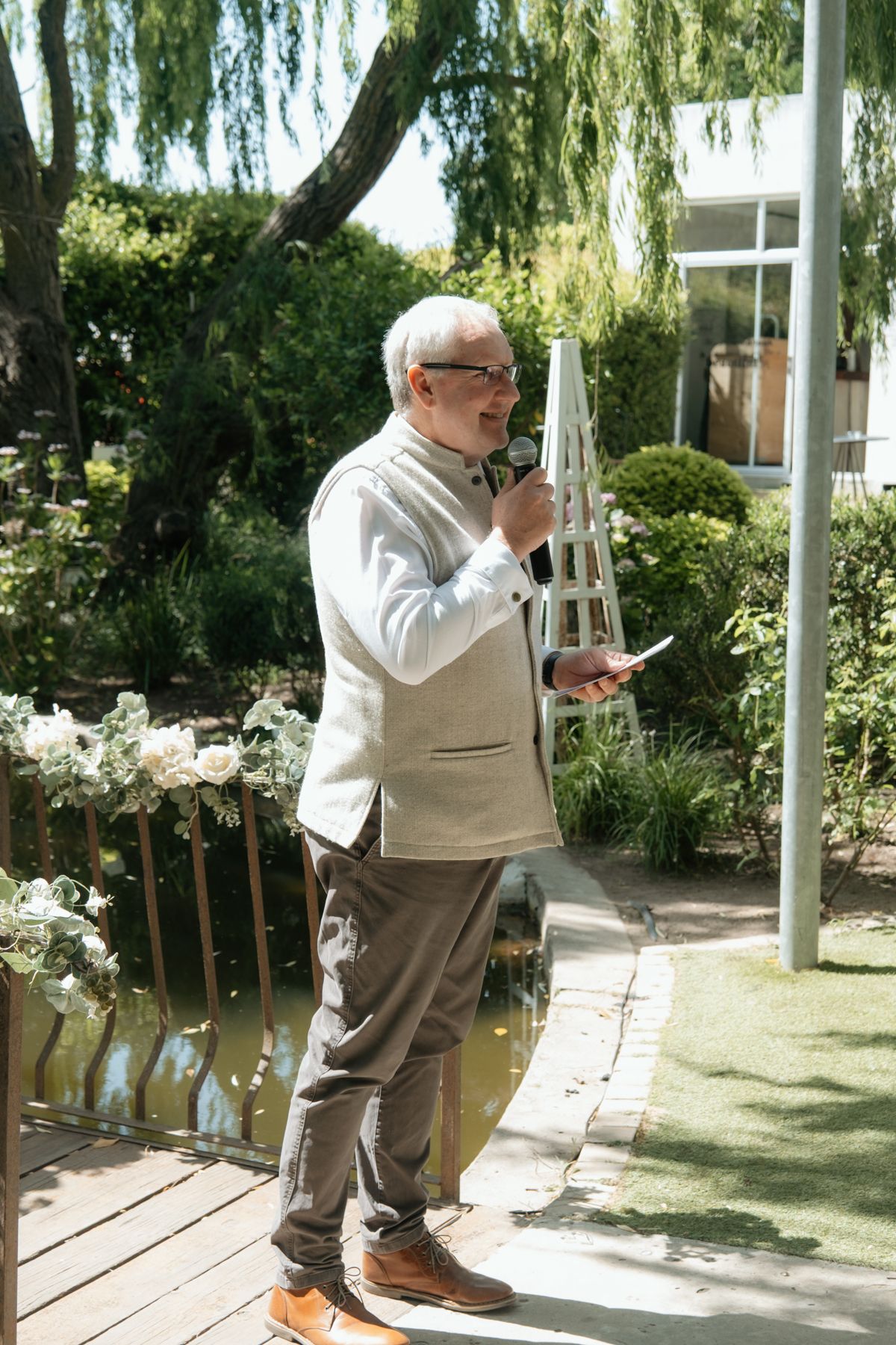 Elderly man in light blazer speaks into microphone at outdoor wedding ceremony by pond.