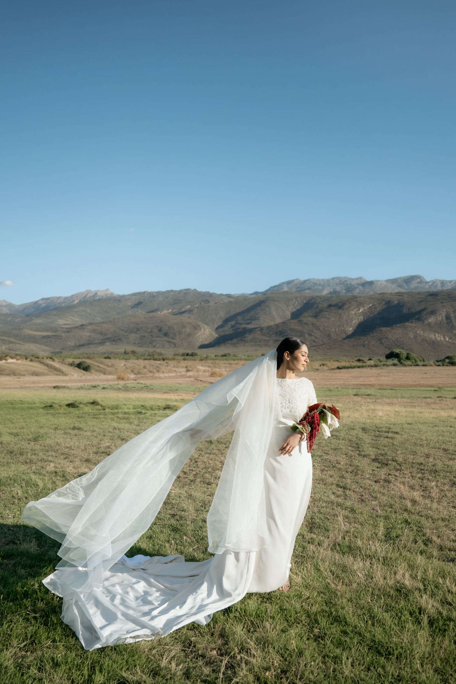 Bride in white gown with flowing veil holds bouquet in open field beneath clear blue mountain sky.