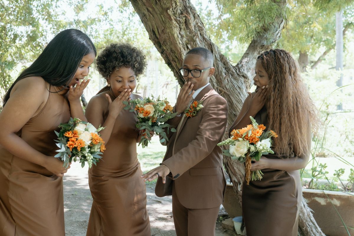 Groom in brown suit shares a joyful moment with three bridesmaids in matching brown dresses holding orange bouquets.