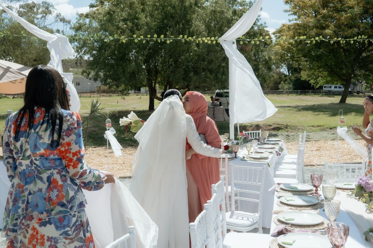 Bride in white gown and veil being guided to her seat at an outdoor wedding reception with white decor.