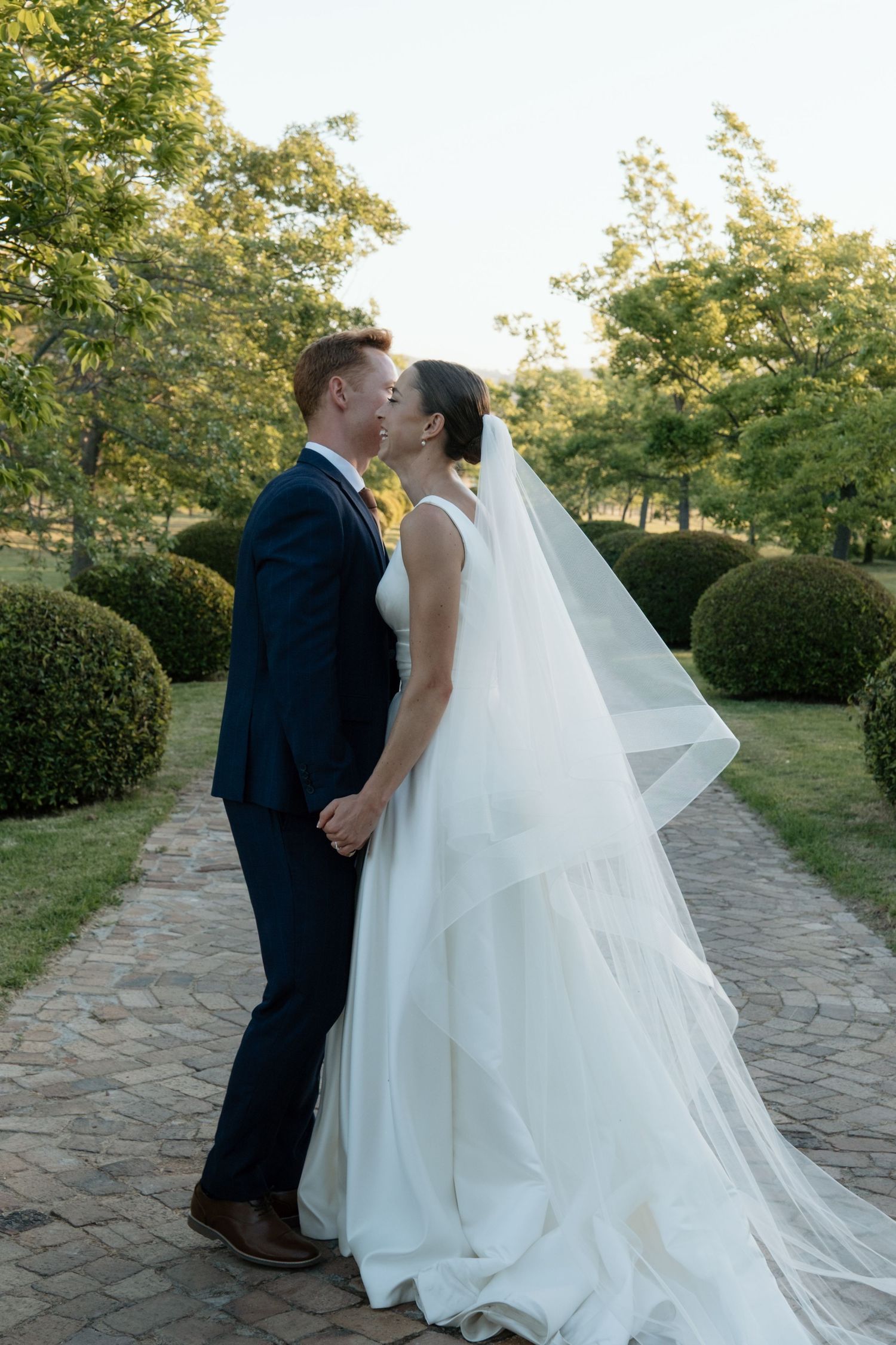 Bride and groom sharing a kiss in a manicured garden, bride wearing a layered cathedral veil.