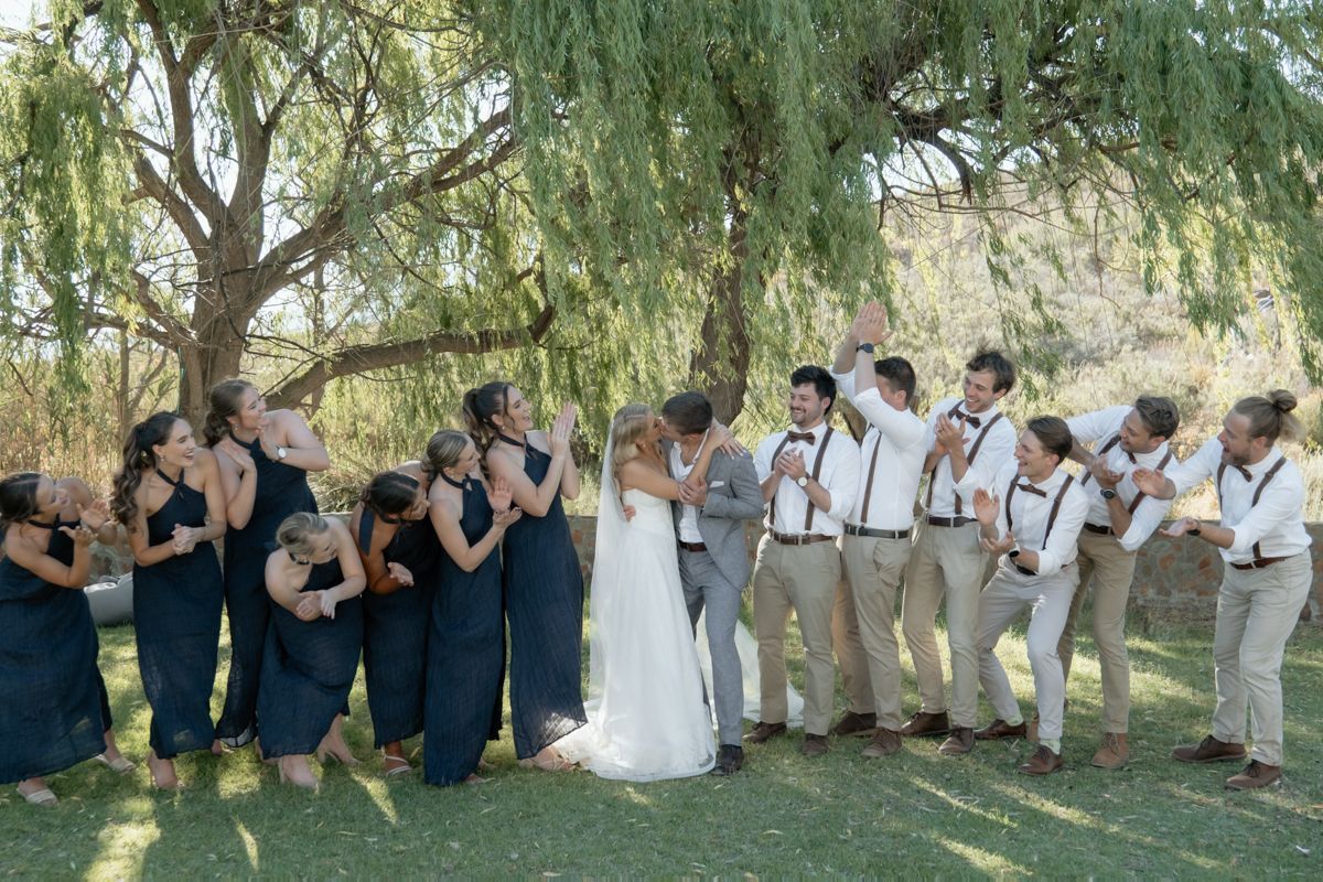 Wedding party stands under willow tree, bridesmaids in navy dresses and groomsmen in suspenders flank kissing bride and groom.