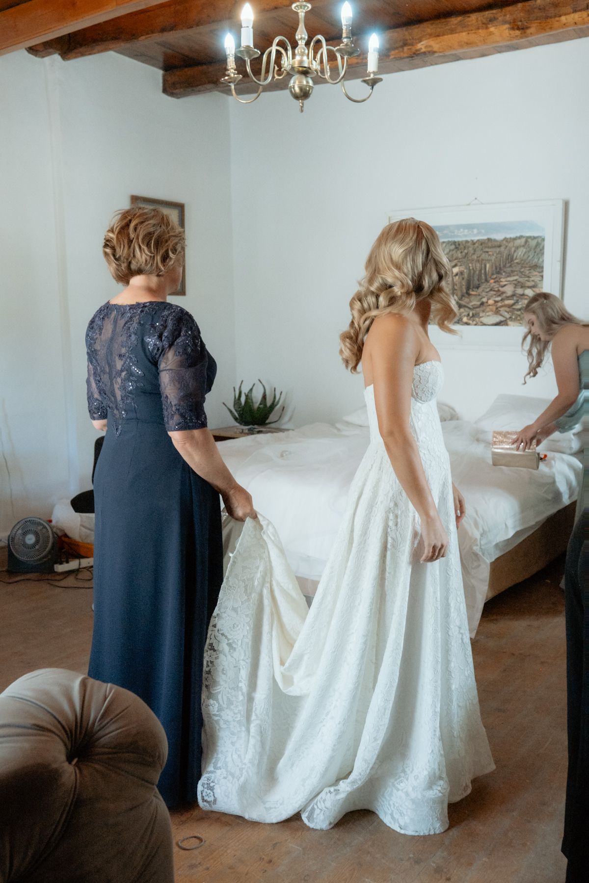 Bride in white lace gown assisted by mother in navy dress during wedding preparations in rustic bedroom.