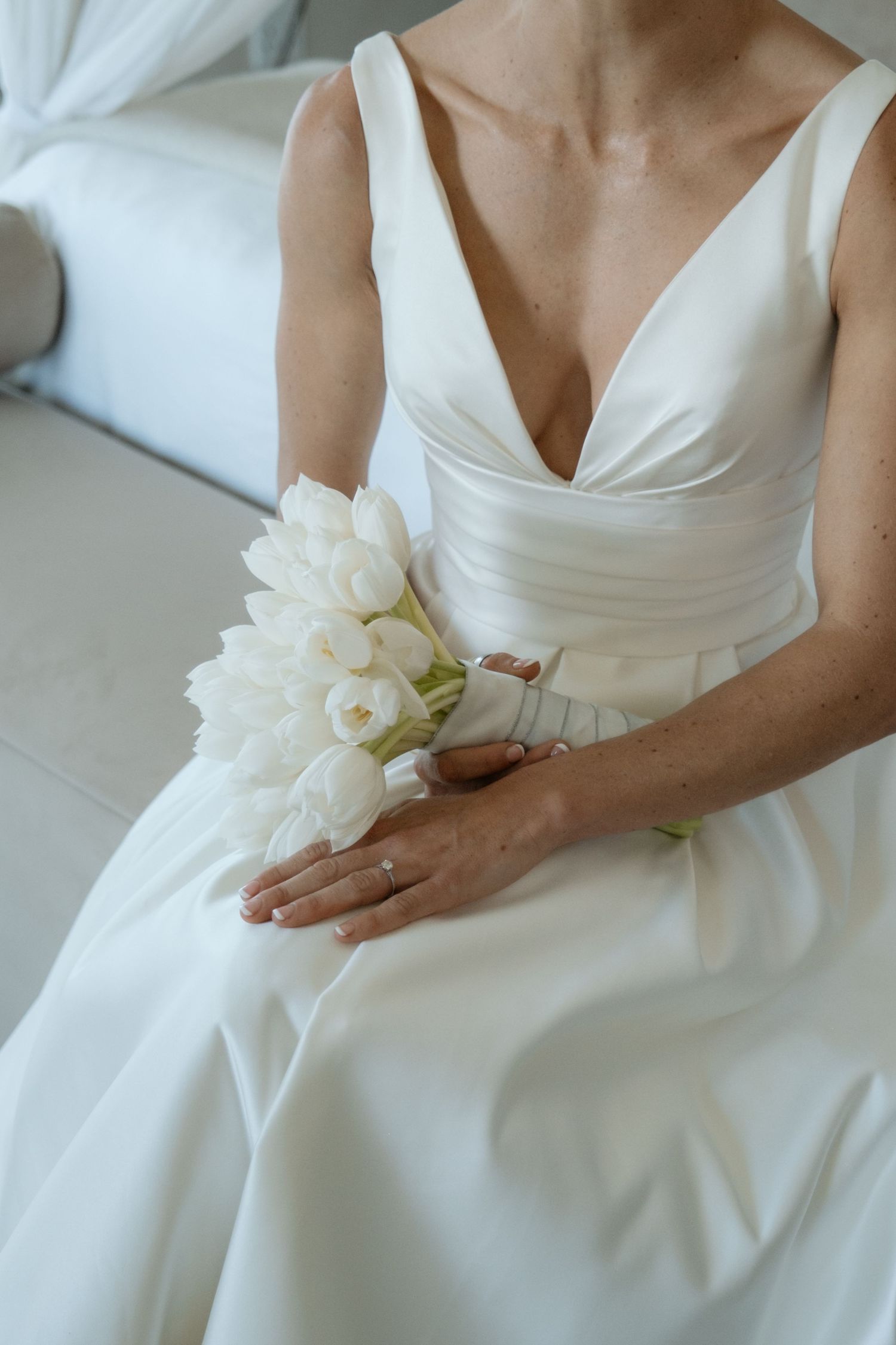 Bride in elegant white V-neck wedding gown holding a bouquet of white tulips while seated.