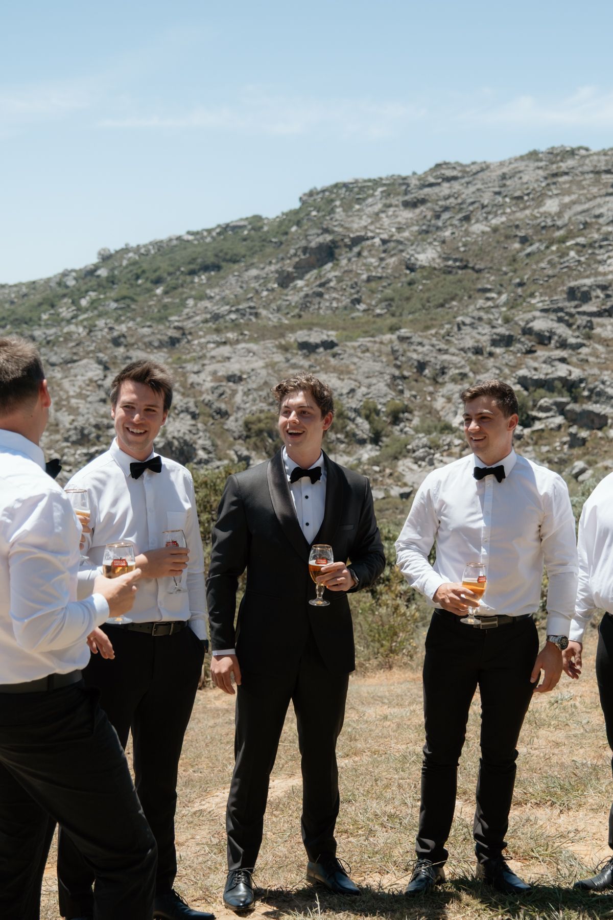 Groomsmen in black tuxedos and bow ties celebrating with champagne glasses outdoors near a rocky mountain backdrop.
