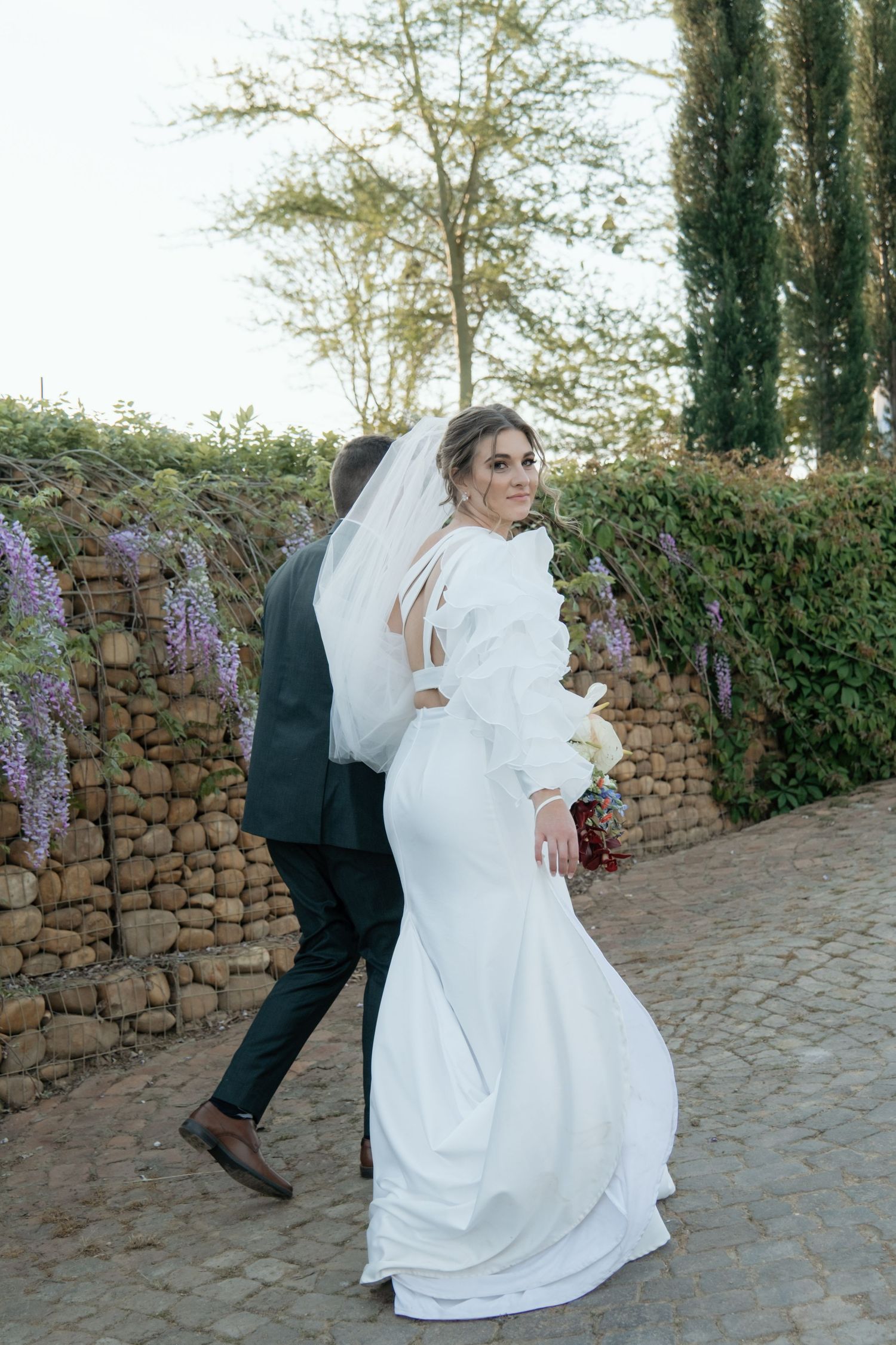 Couple in wedding attire posing against stone wall with purple flowers in Stellenbosch