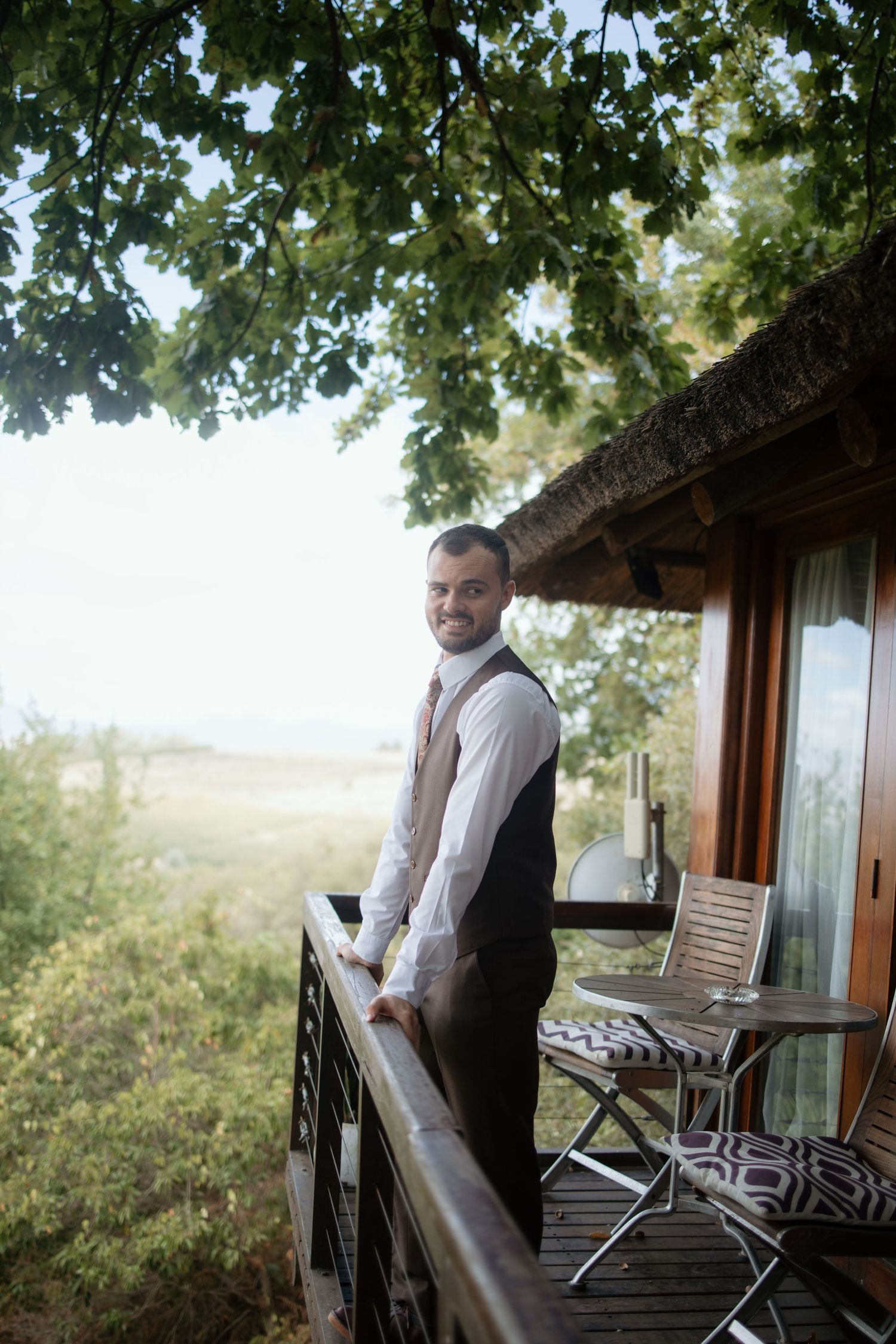 A smiling groom in a vest stands on a rustic balcony surrounded by lush greenery and scenic countryside views.