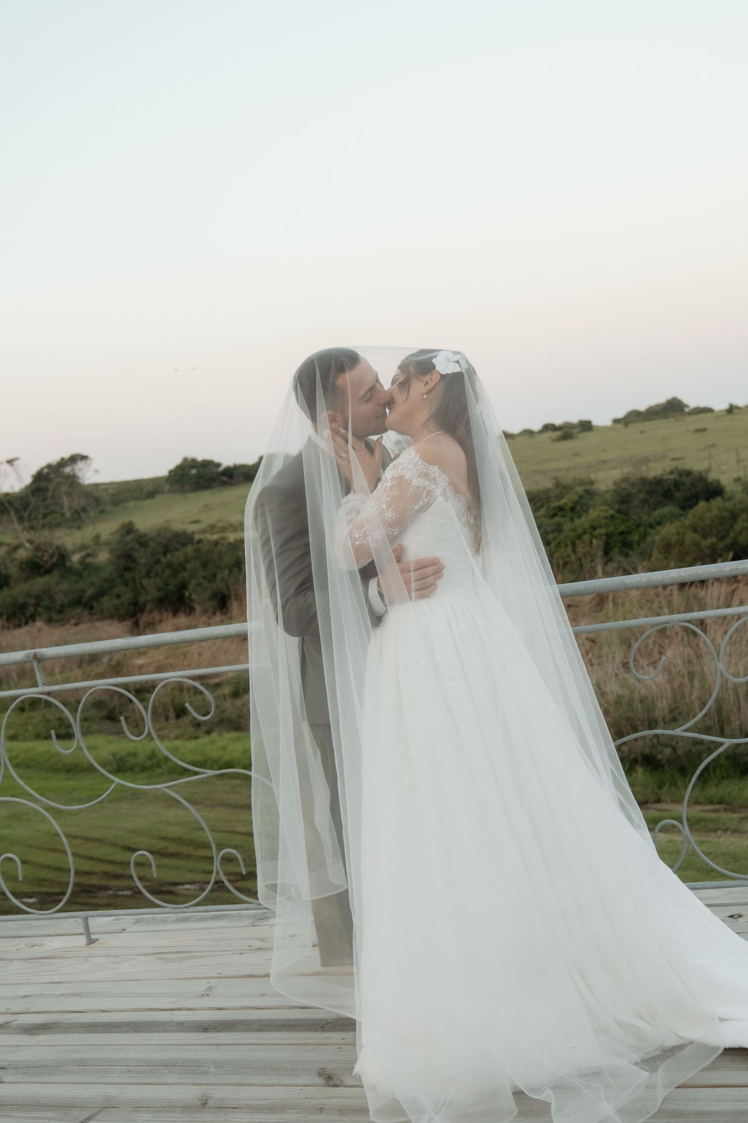 Bride and groom kissing under flowing veil on outdoor terrace with green landscape at twilight.
