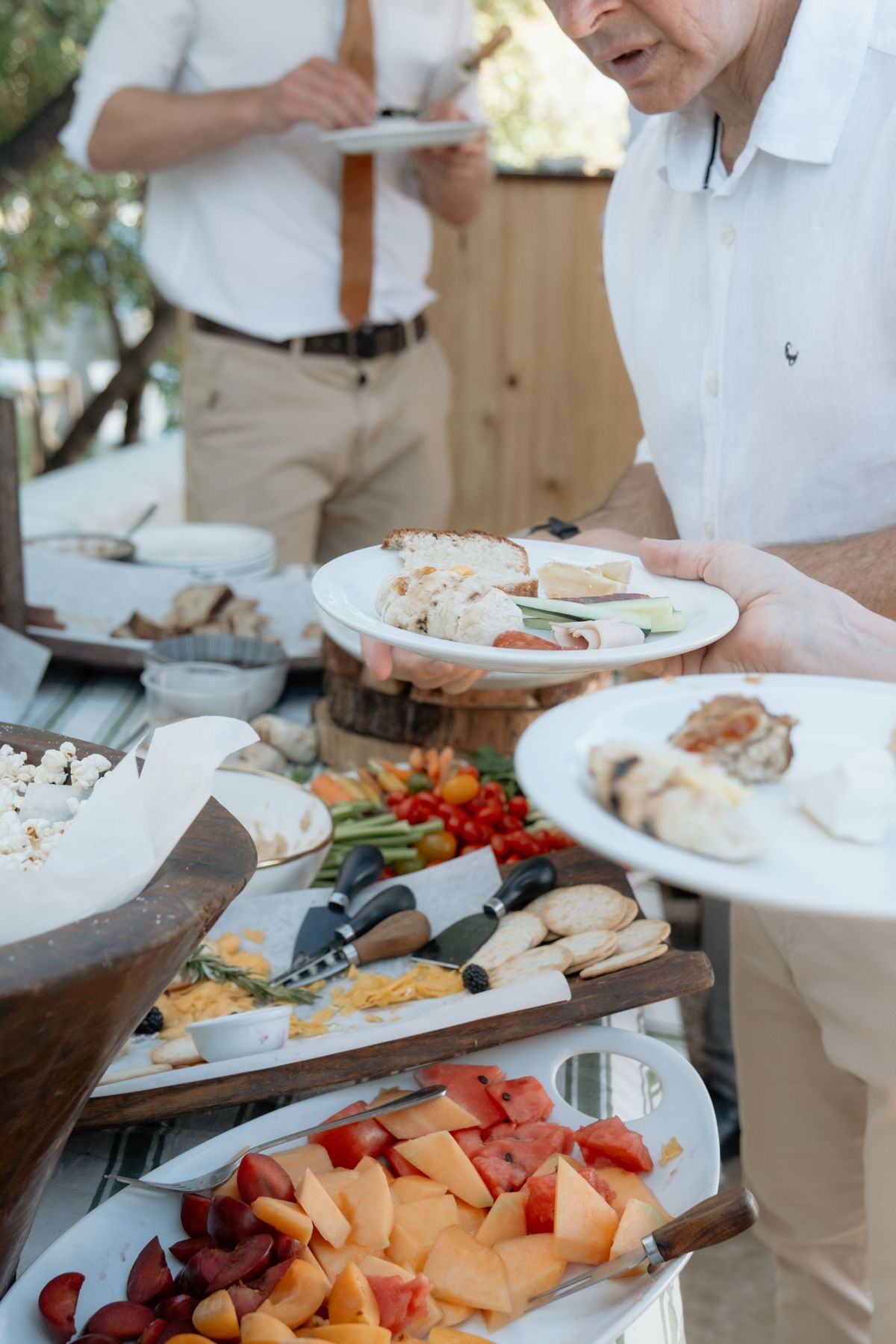 Guests serving themselves at an elegant outdoor wedding buffet featuring fresh fruits, vegetables, and appetizers on tiered wooden displays.