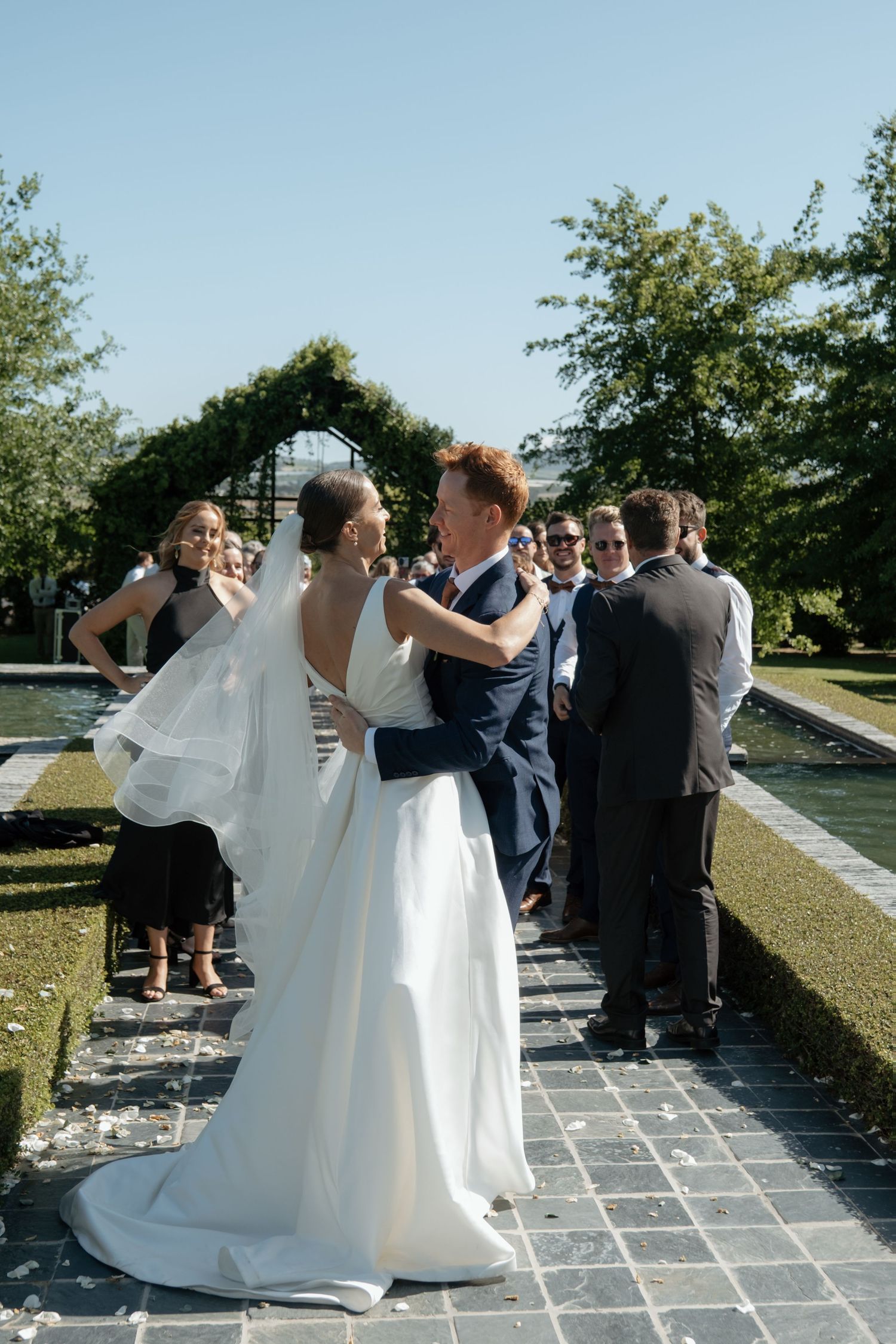 Bride and groom share a romantic moment on a garden pathway surrounded by wedding guests at an outdoor ceremony.