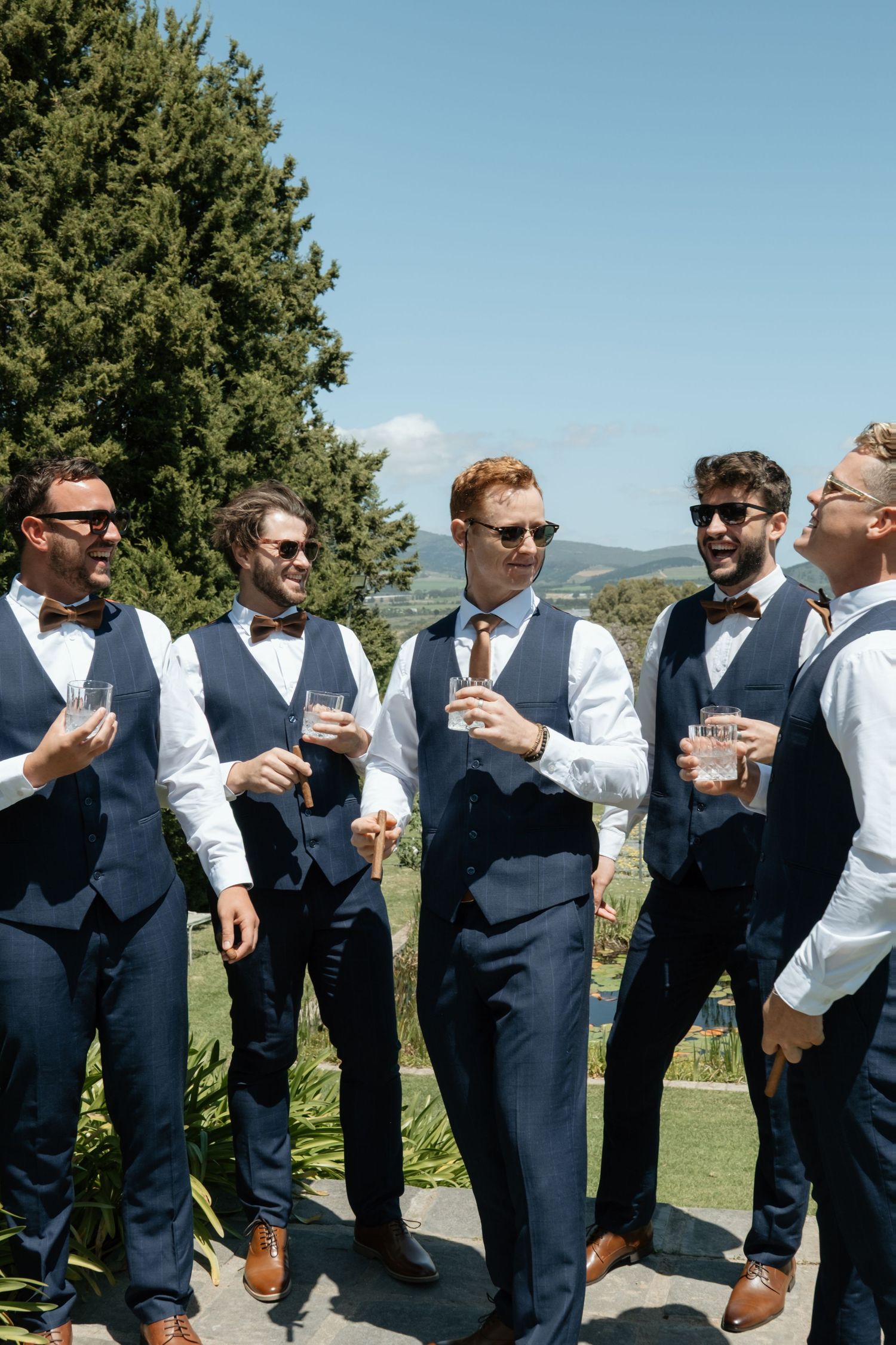 Five groomsmen in navy suits and bow ties holding drinks outdoors on a sunny wedding day.