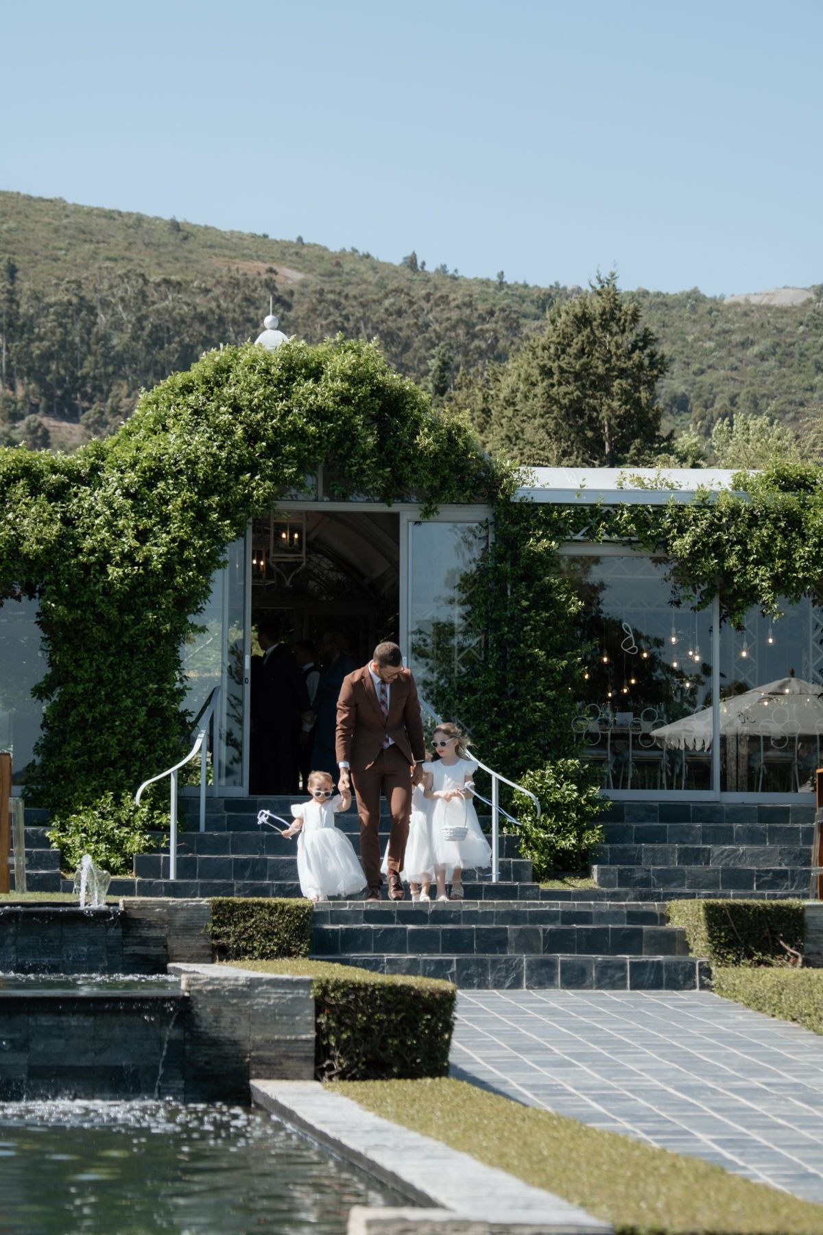 Man in brown suit escorts two young flower girls in white dresses down steps at a lush garden venue.