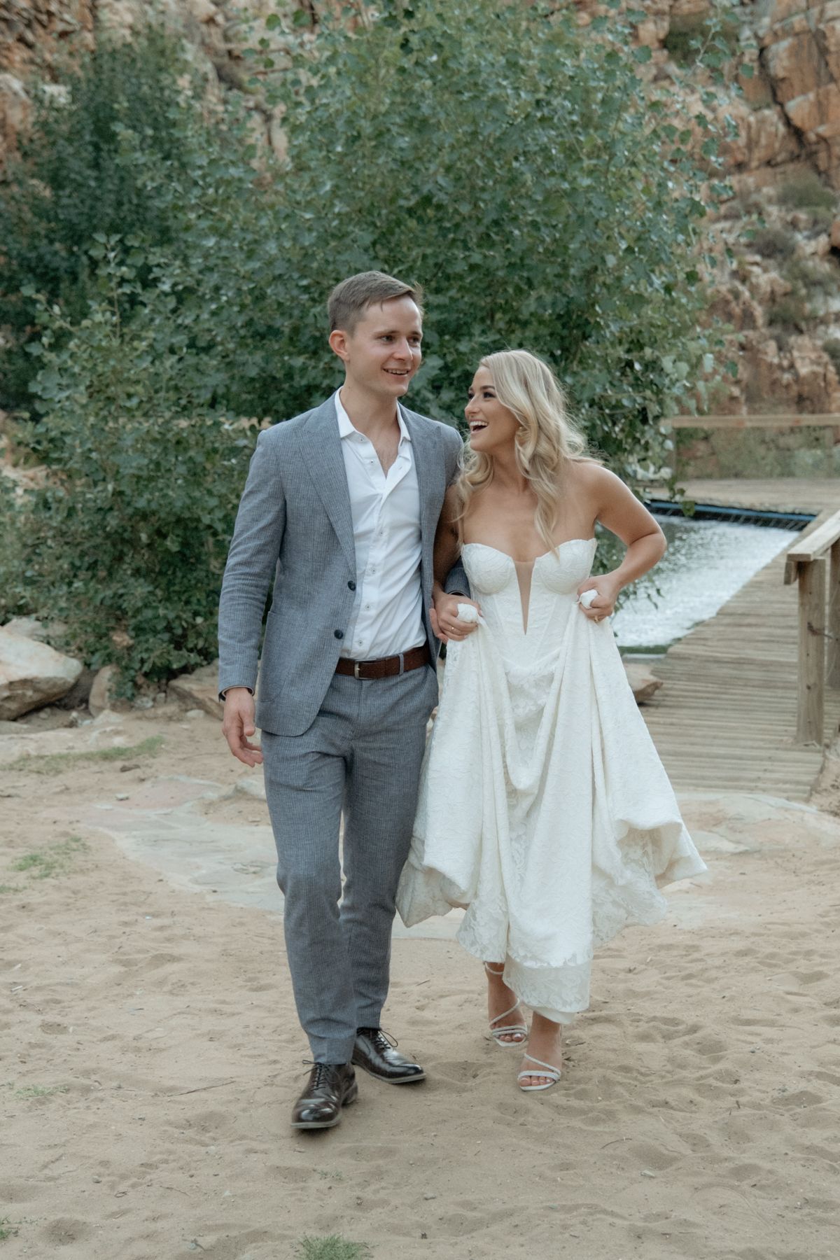 Smiling bride and groom walking together on sandy path near pool in relaxed outdoor wedding setting.