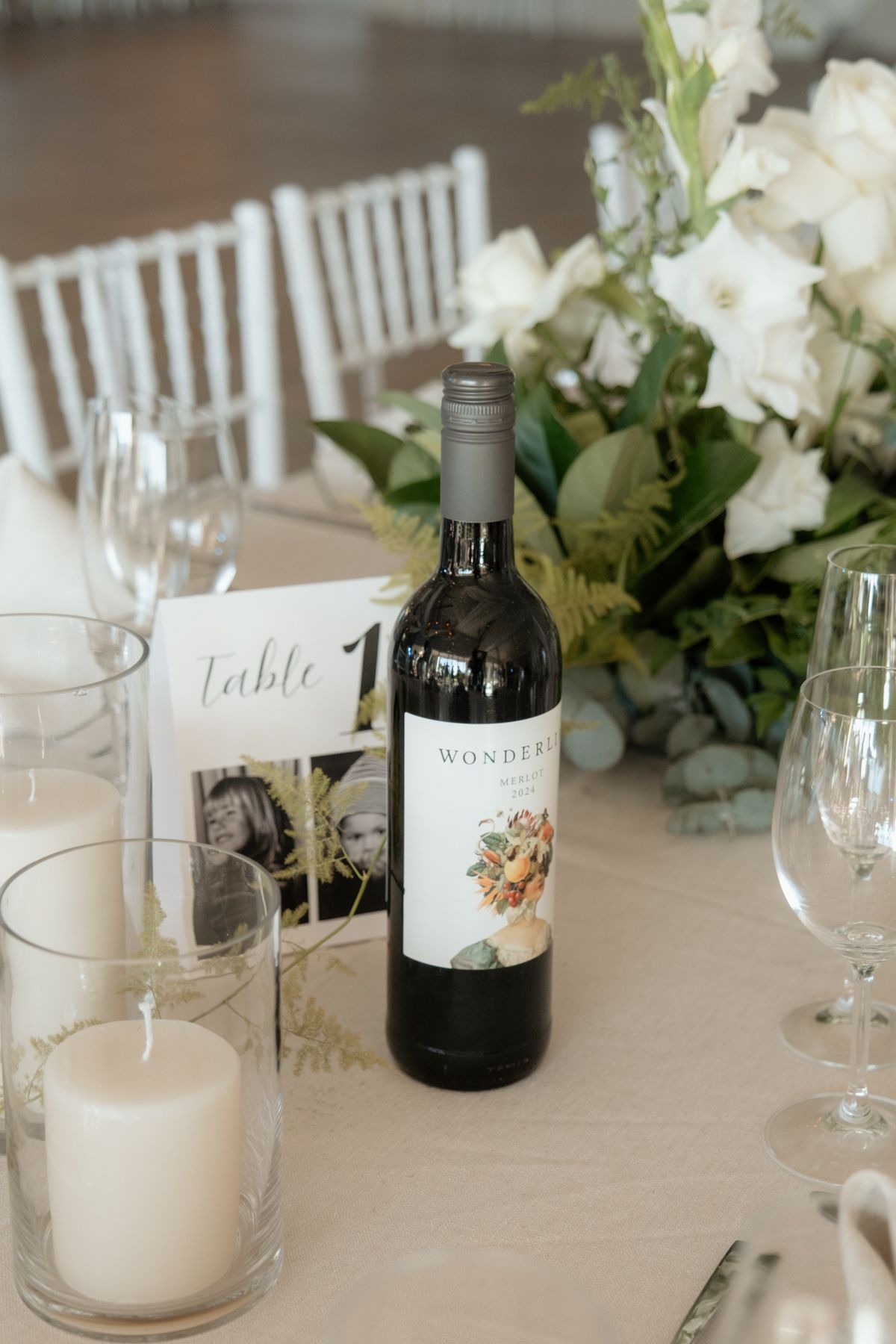 Elegant wedding reception table with wine bottle, white floral centerpiece, candles, and table number card.