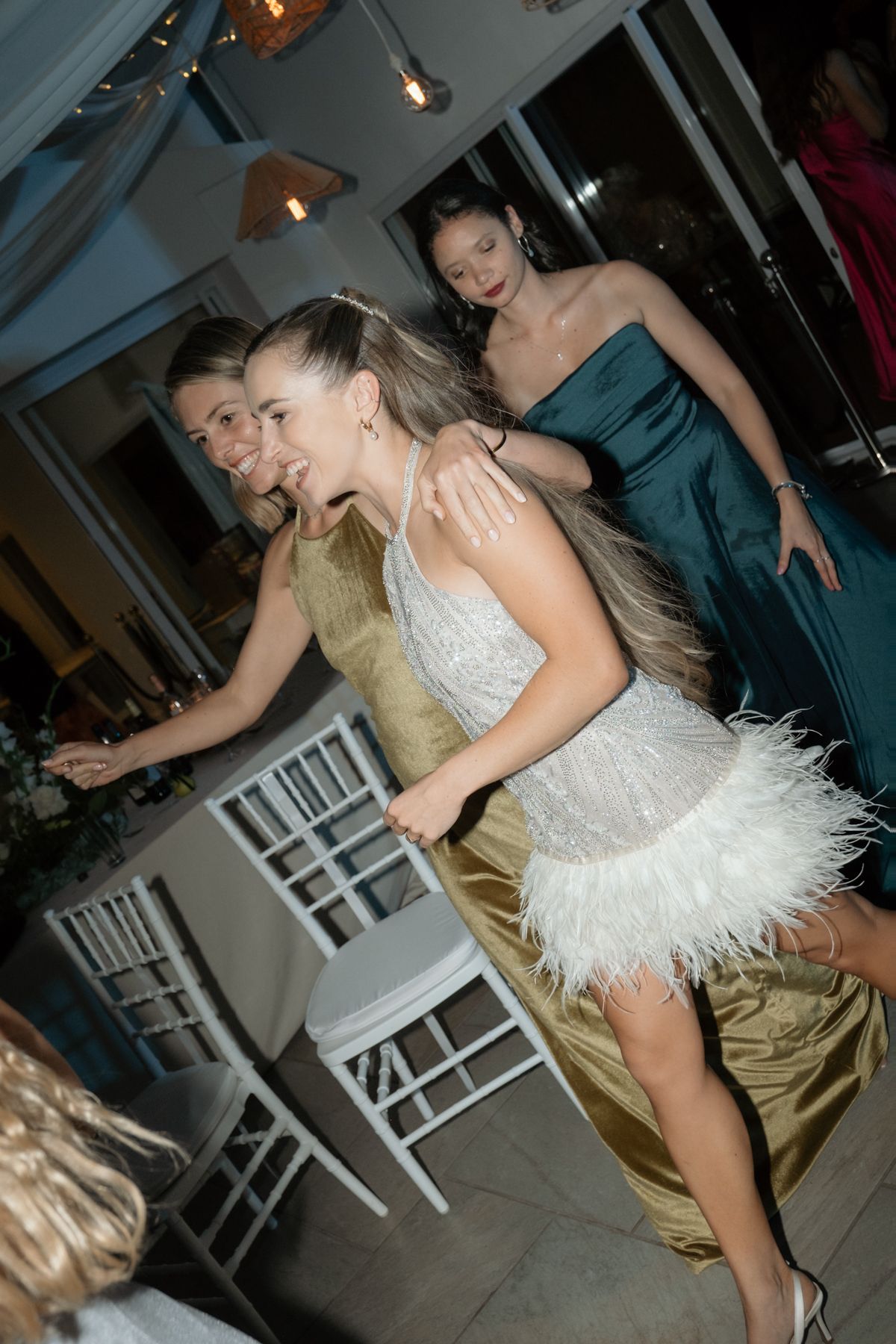 Bride in a white feathered mini dress dancing energetically with guests at a wedding reception.