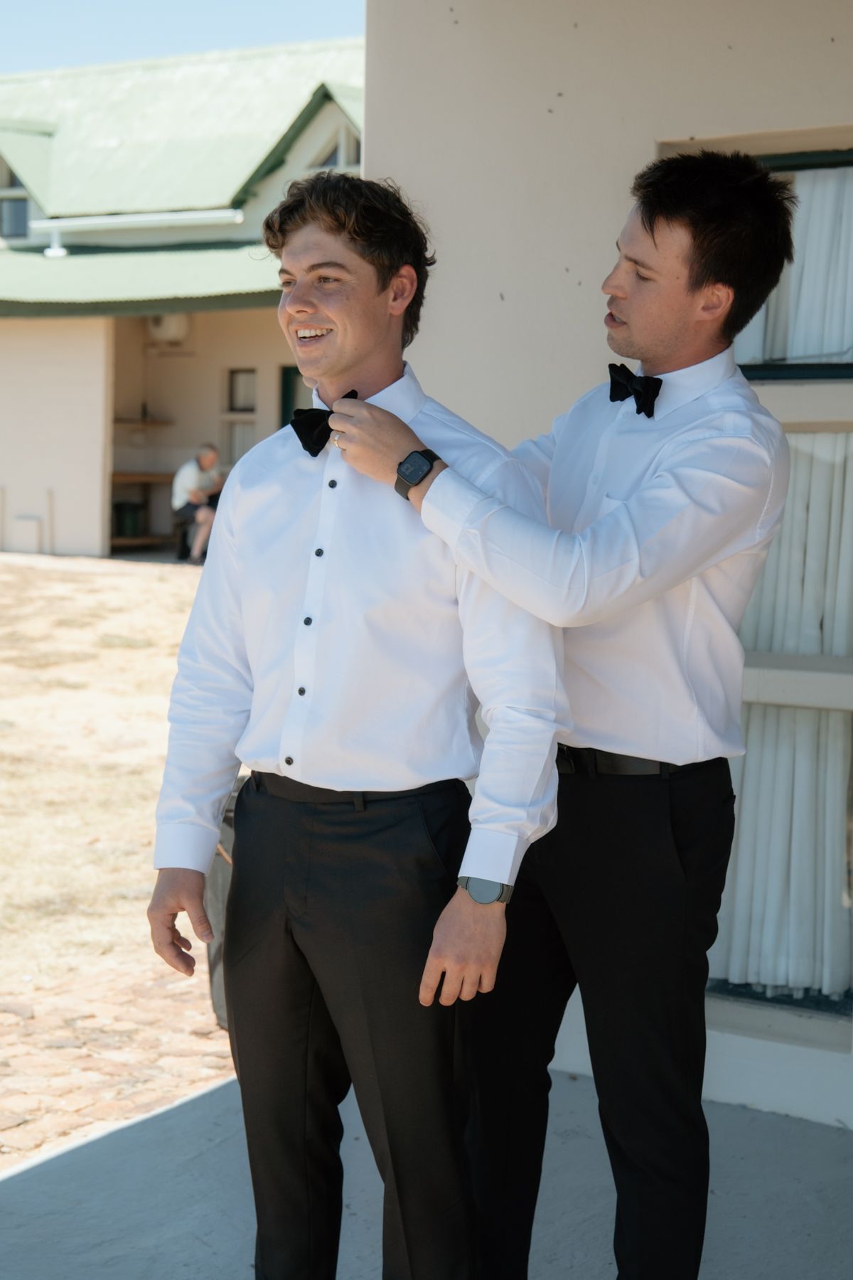 Smiling groom having bow tie adjusted by groomsman outside venue in matching white shirts and black trousers.