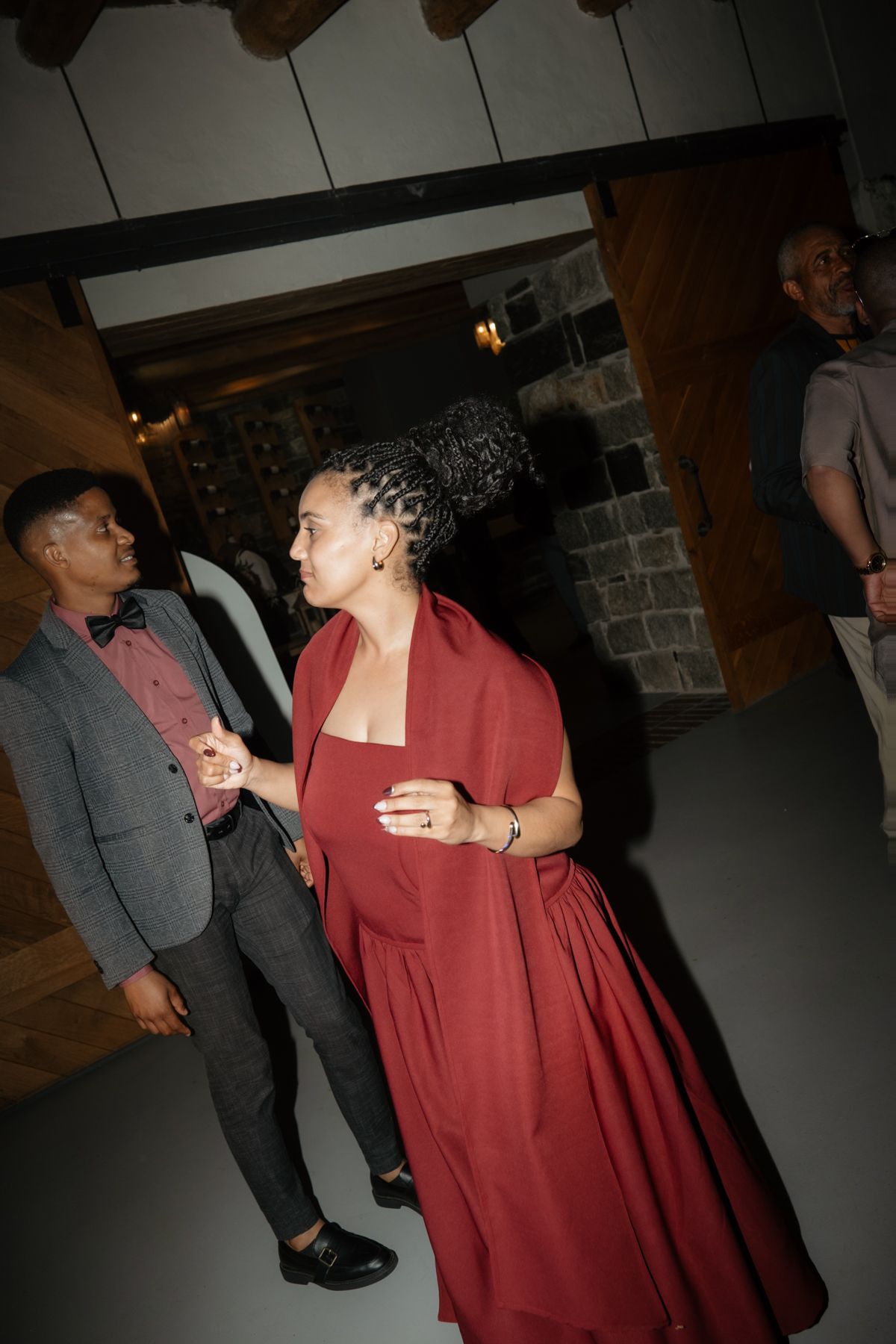Wedding guest in flowing red dress and gray-suited guest mingle near rustic stone doorway at reception venue.