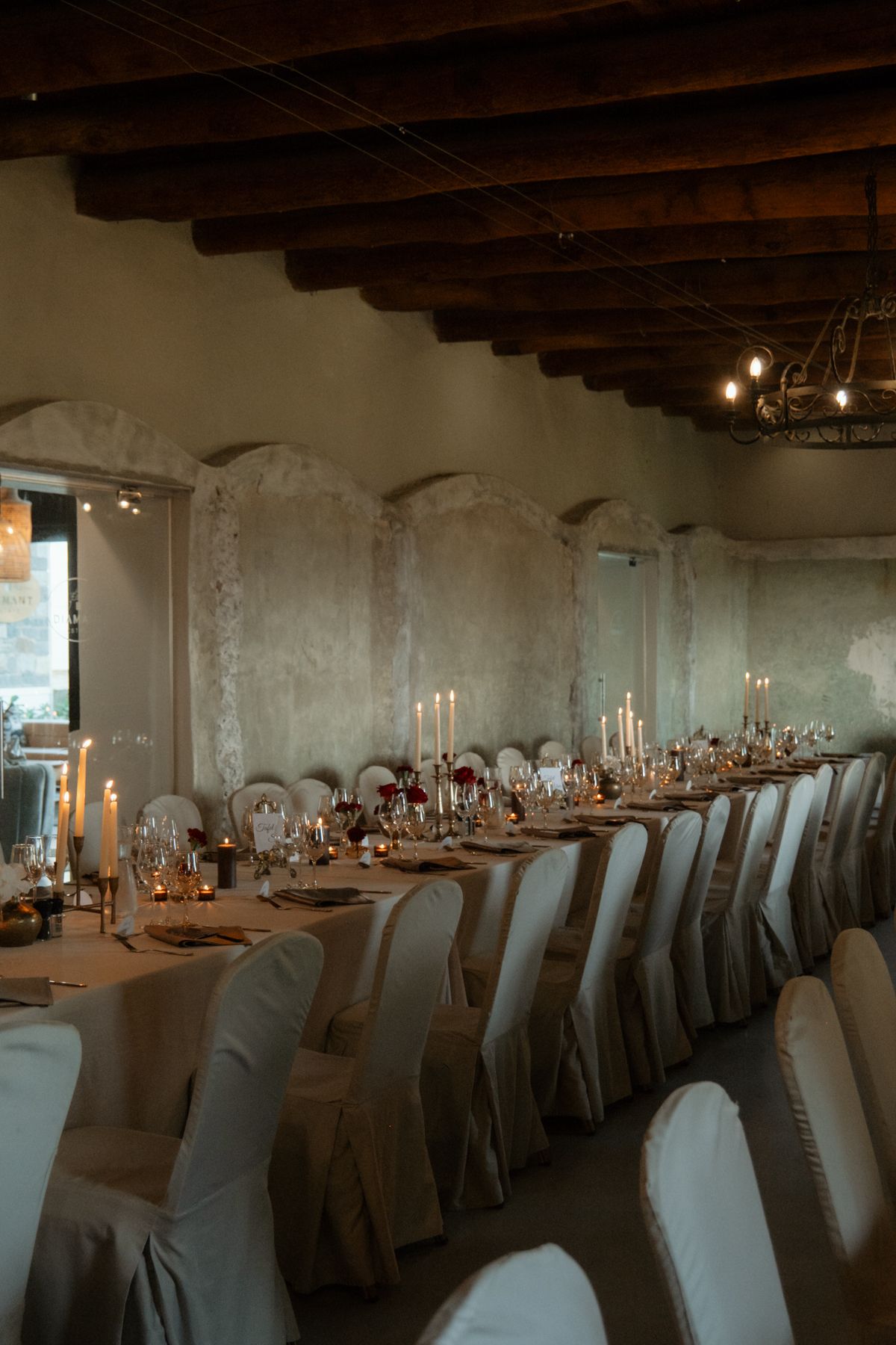 Elegant wedding reception hall with long candlelit table, white chair covers, and rustic wooden ceiling beams.
