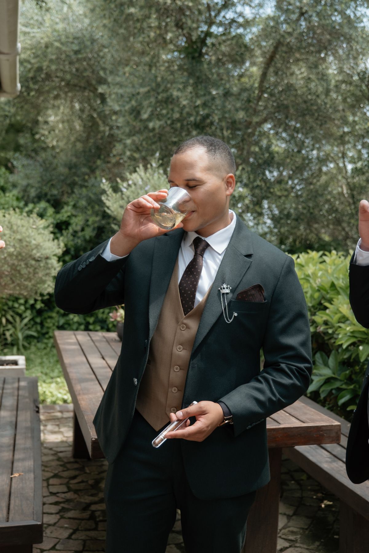 Dapper groom in dark green suit drinking champagne outdoors before wedding ceremony.