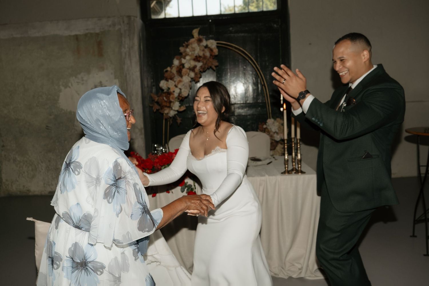 Groom applauds as bride kneels to greet elderly woman in blue hijab during wedding celebration.