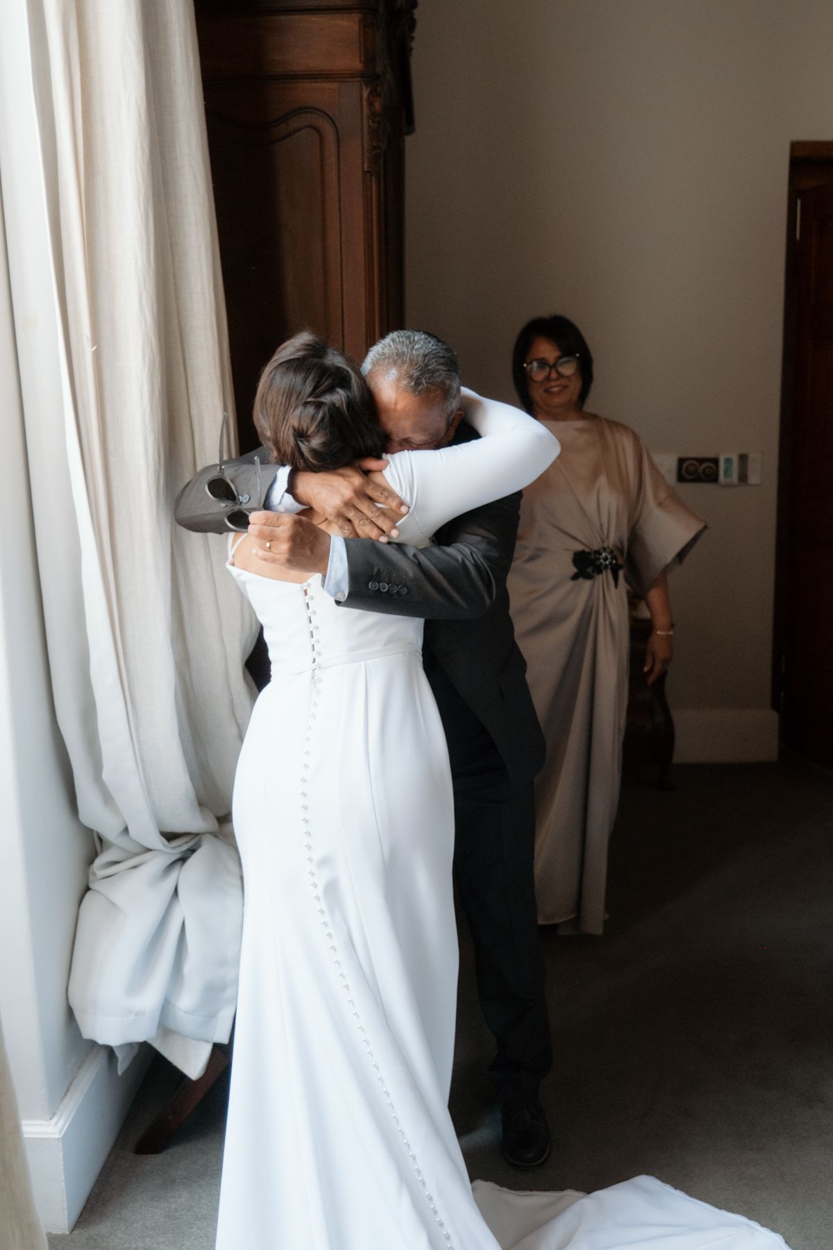 Bride in white gown sharing an emotional hug with a guest while another woman watches warmly.