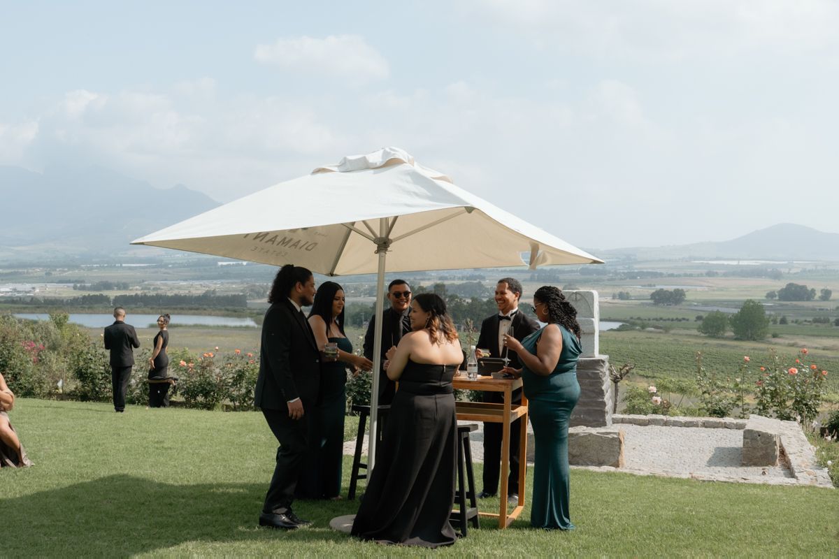Wedding guests gather under a white umbrella on a scenic vineyard lawn with mountain views in the background.