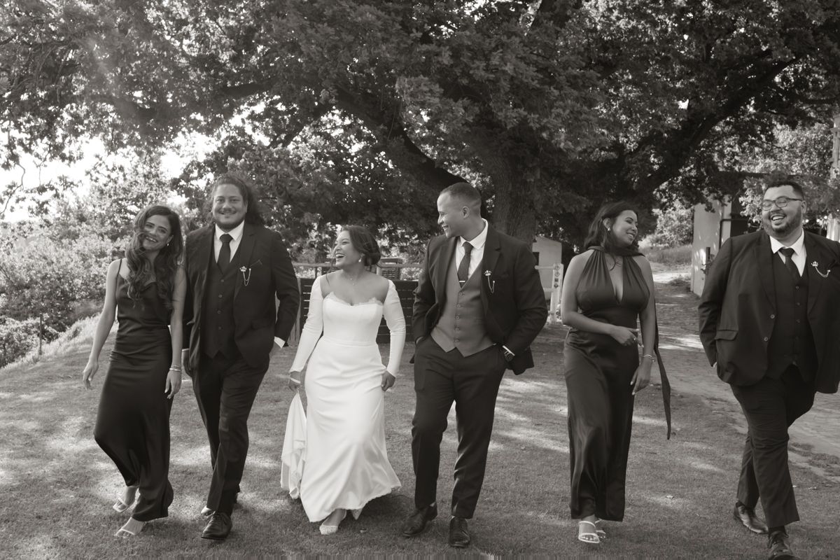Black and white photo of bride, groom, and wedding party walking together under large oak tree.