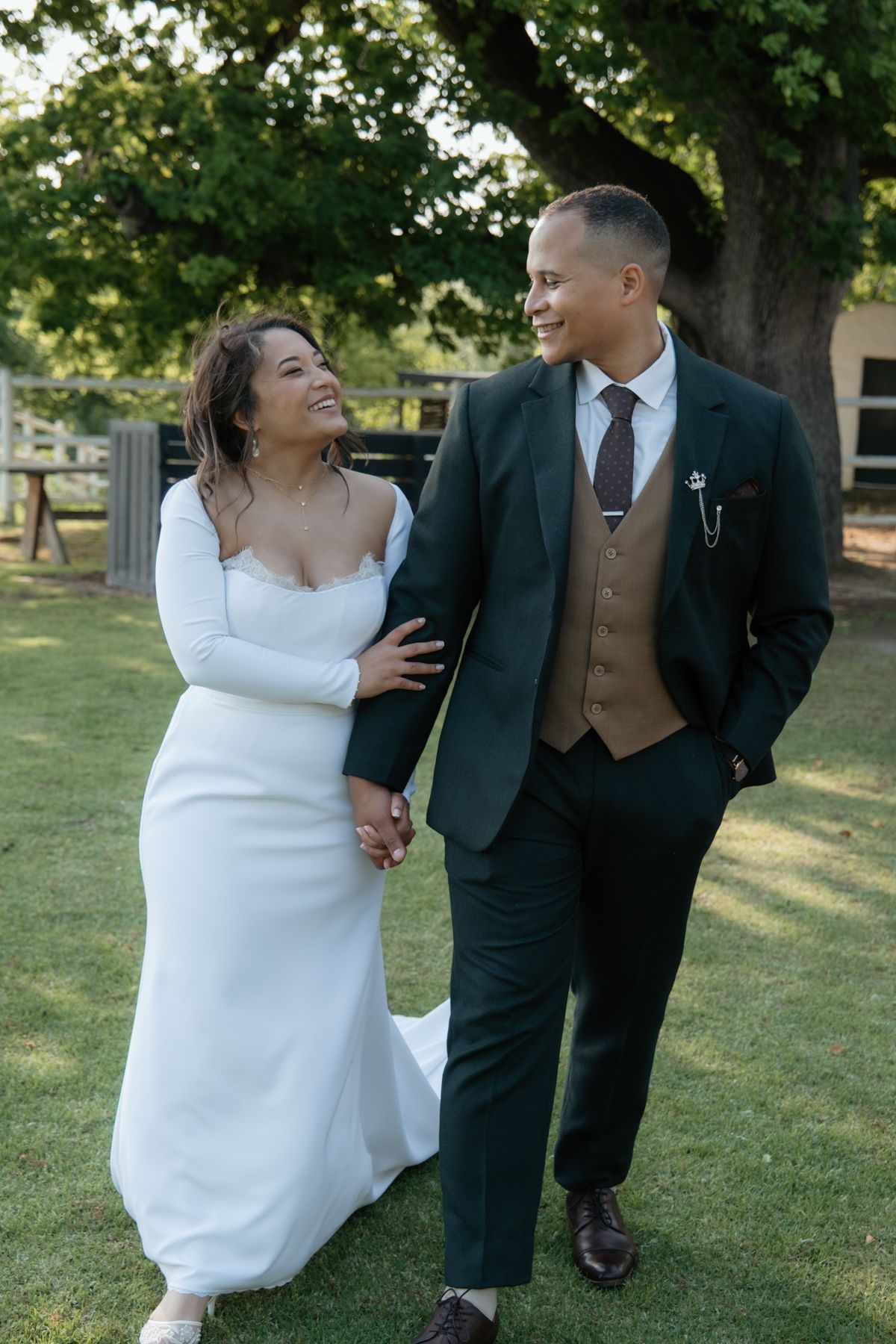 Smiling bride and groom walking hand in hand on green lawn beside a large tree.