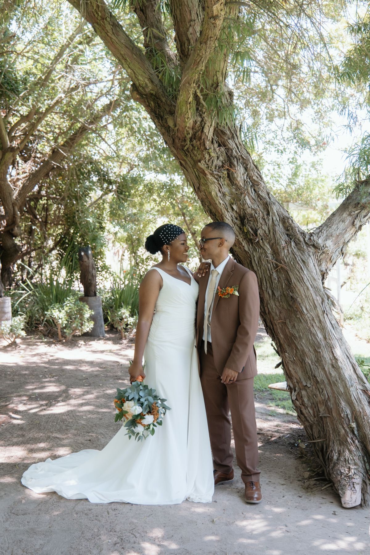 Black bride and groom share tender moment leaning against large tree, bride holding white and orange bouquet.