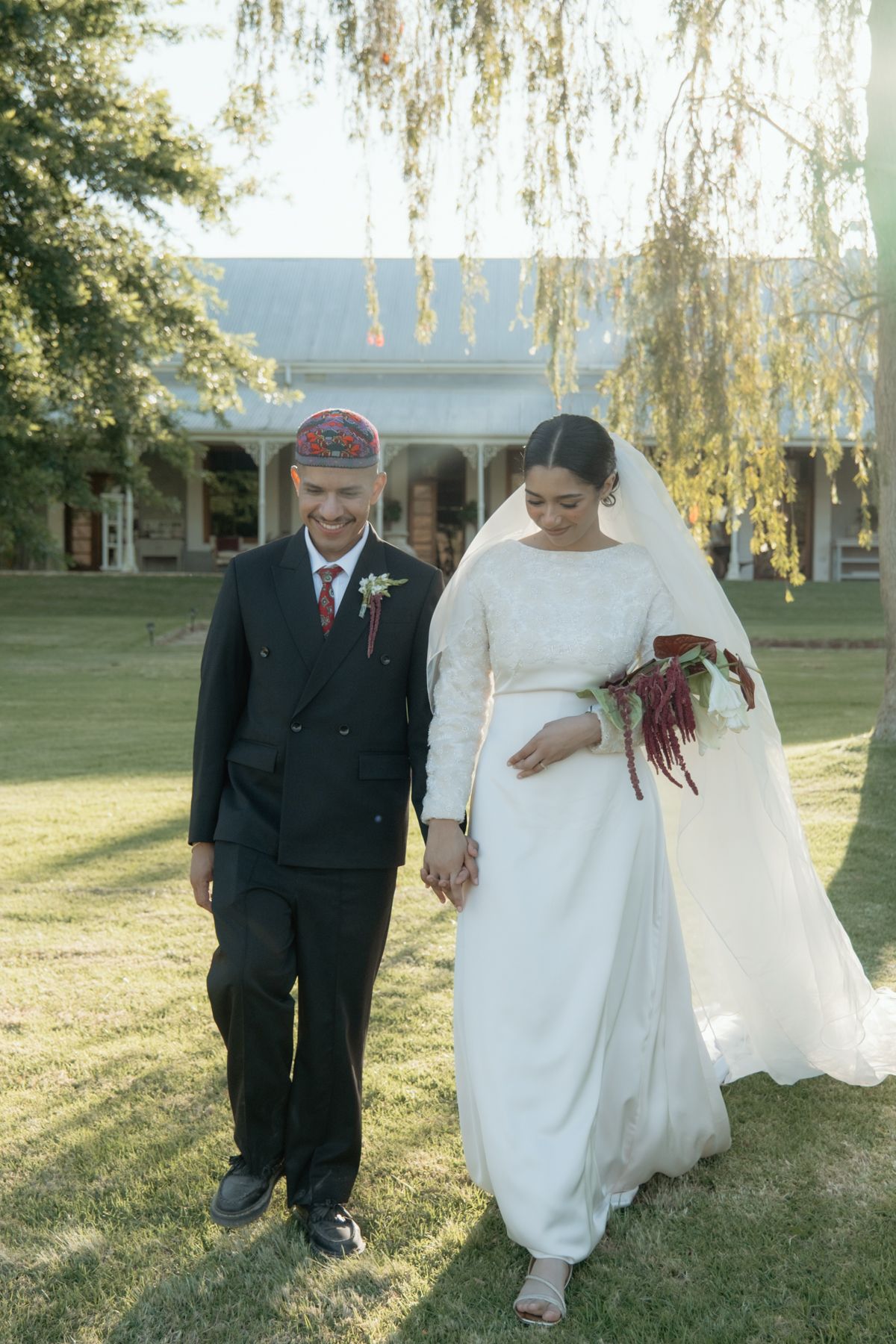 Bride in white gown and groom in black suit with embroidered cap walking together at outdoor wedding.