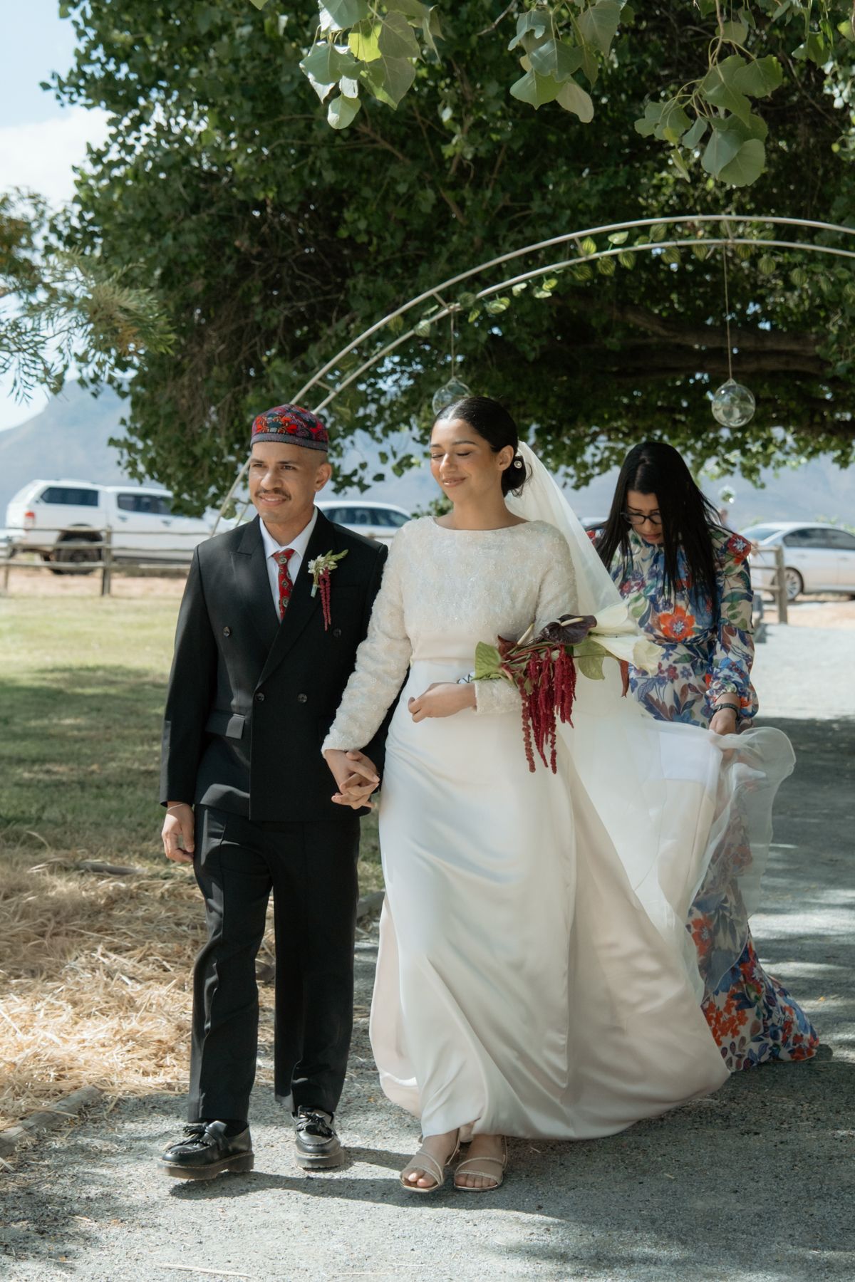 Bride in white gown walks with groom in embroidered cap under floral arch, assisted by woman in floral dress.