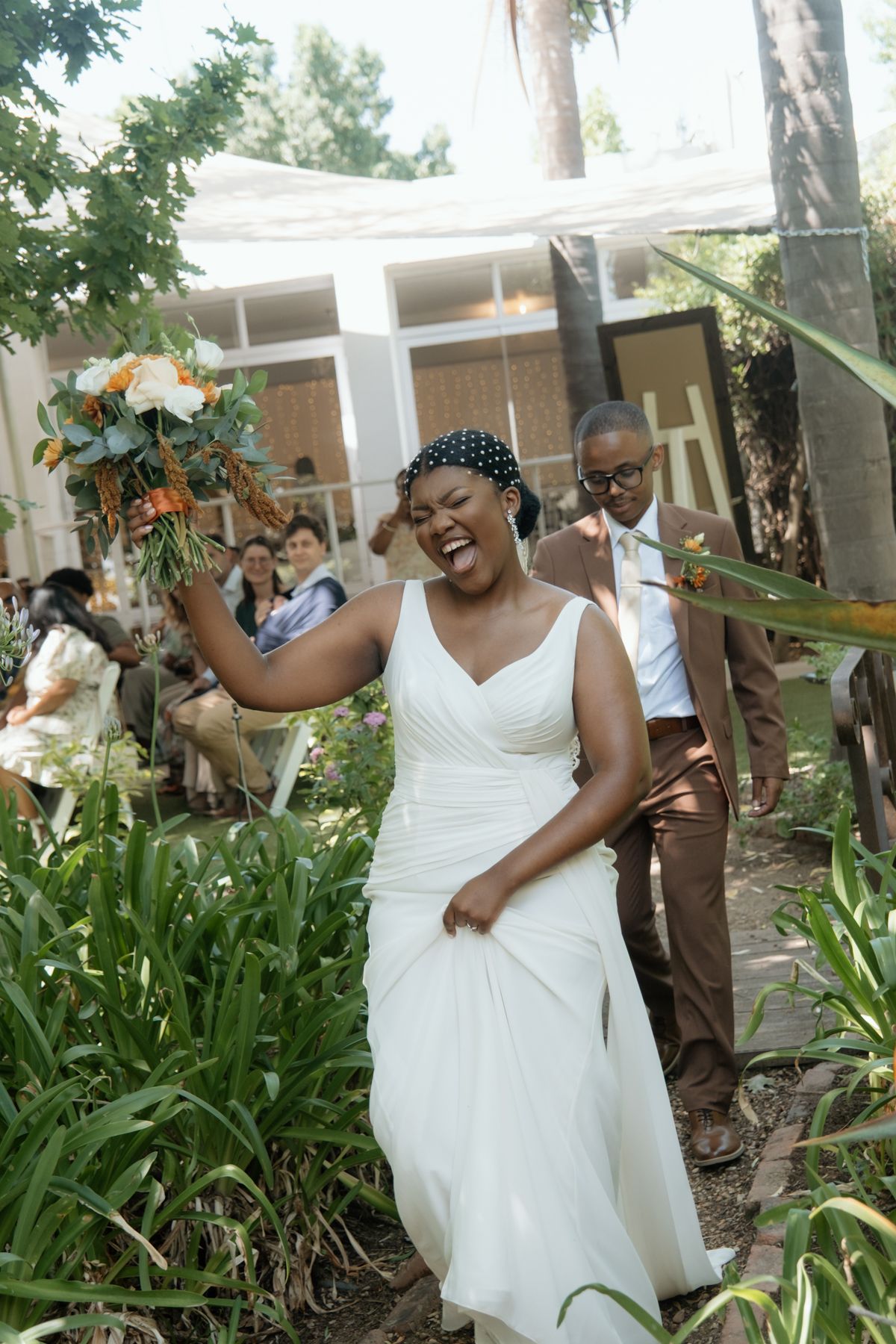 Joyful bride in white gown raises bouquet while laughing as groom follows through garden after ceremony.