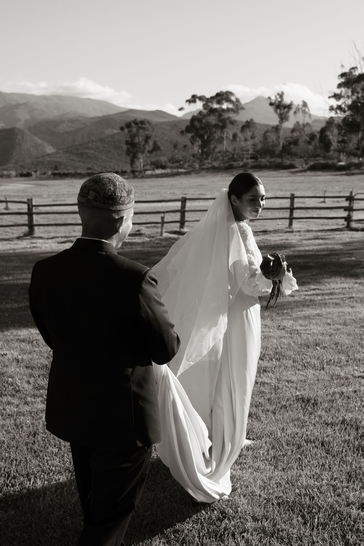 Black and white photo of bride in veil and groom in tuxedo standing on grass with mountain backdrop.