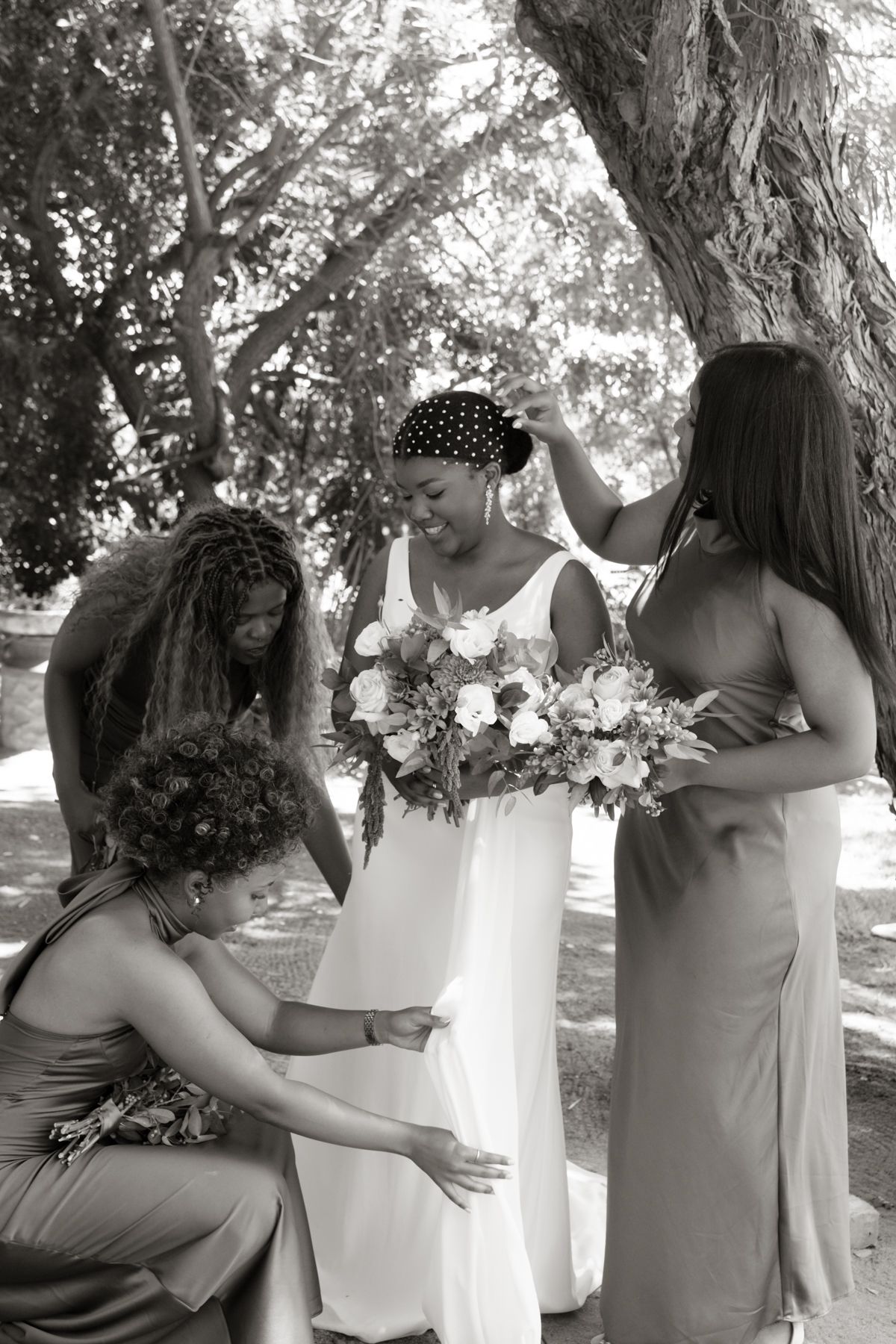 Black and white photo of bridesmaids in brown dresses attending to smiling bride holding bouquet under a tree.