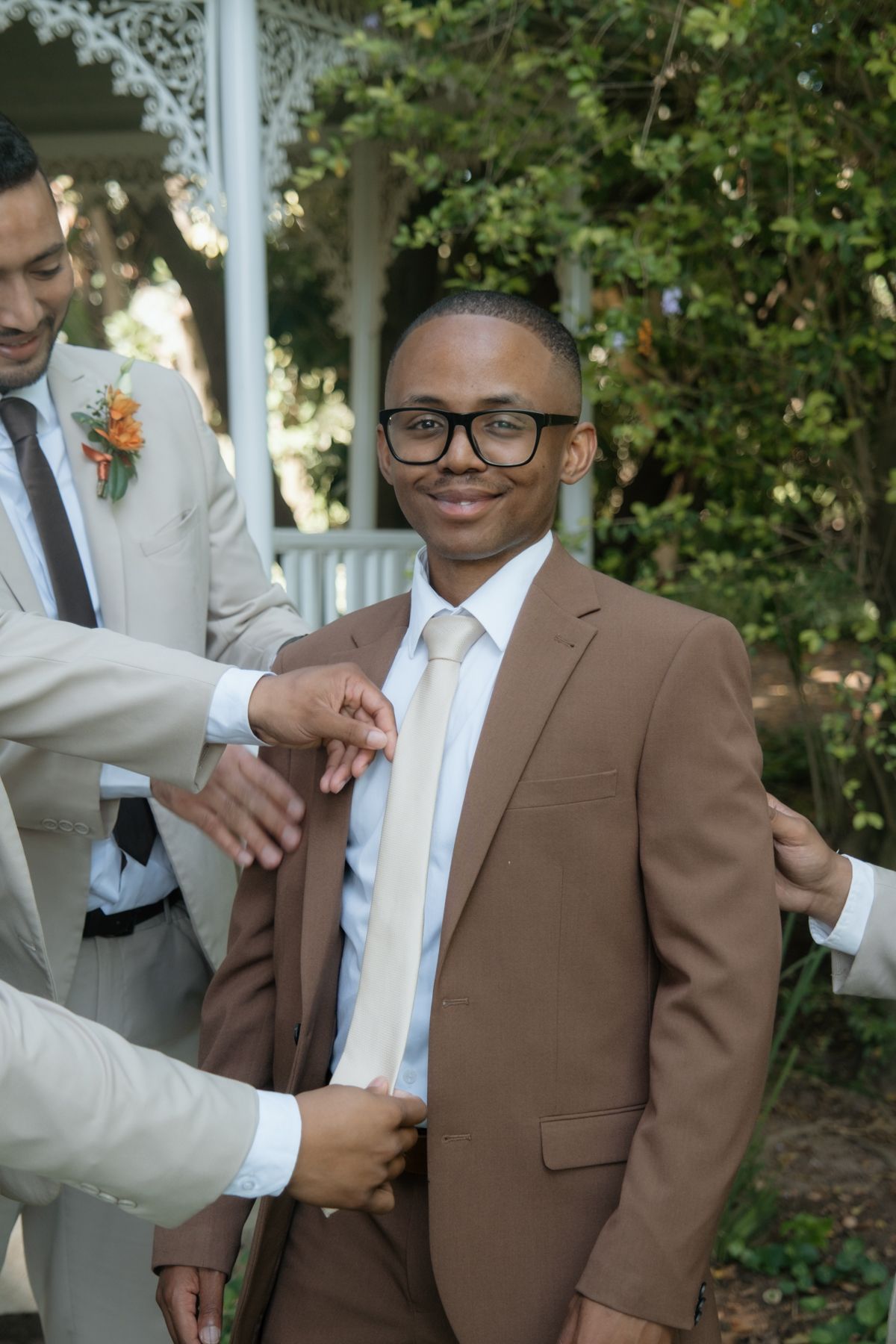 Groomsmen help adjust the brown suit jacket of a smiling young man wearing glasses outdoors.