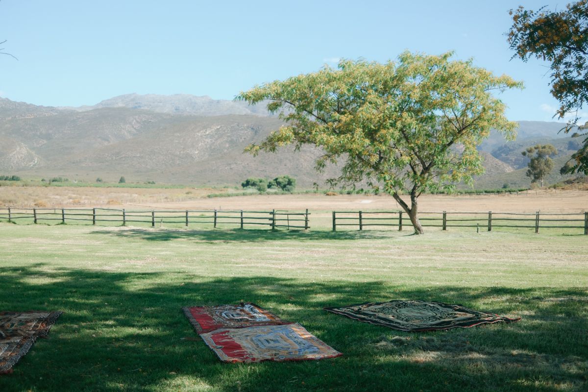 Scenic outdoor wedding venue with colorful rugs on green grass, lone tree, fence, and mountains.