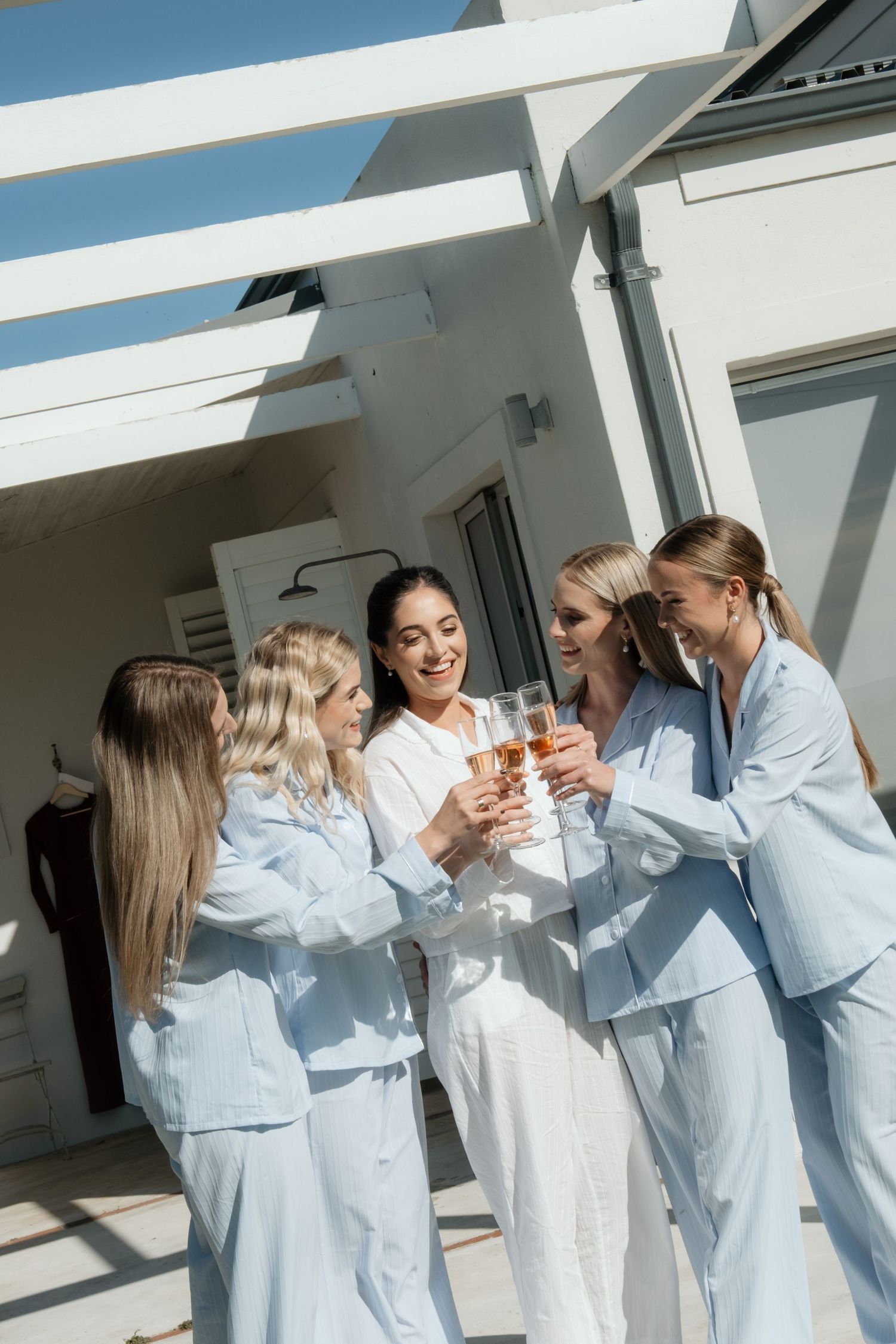 Five women in matching blue pajamas toast champagne glasses together on a sunny outdoor terrace.