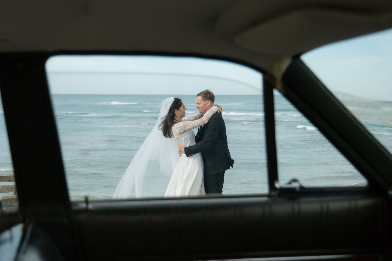 Bride and groom embrace by the ocean, framed through a vintage car window on their wedding day.