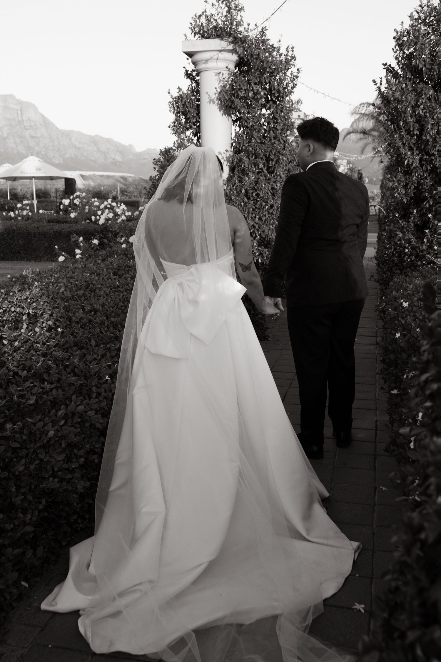 A couple walks along a garden path with the bride wearing a flowing white wedding dress and veil.