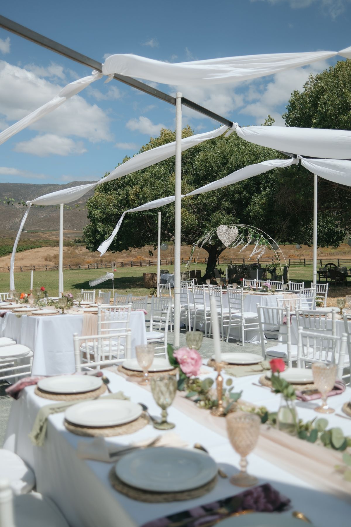Outdoor wedding venue with white draped canopy, elegantly set tables, chairs, and heart-shaped arch backdrop.