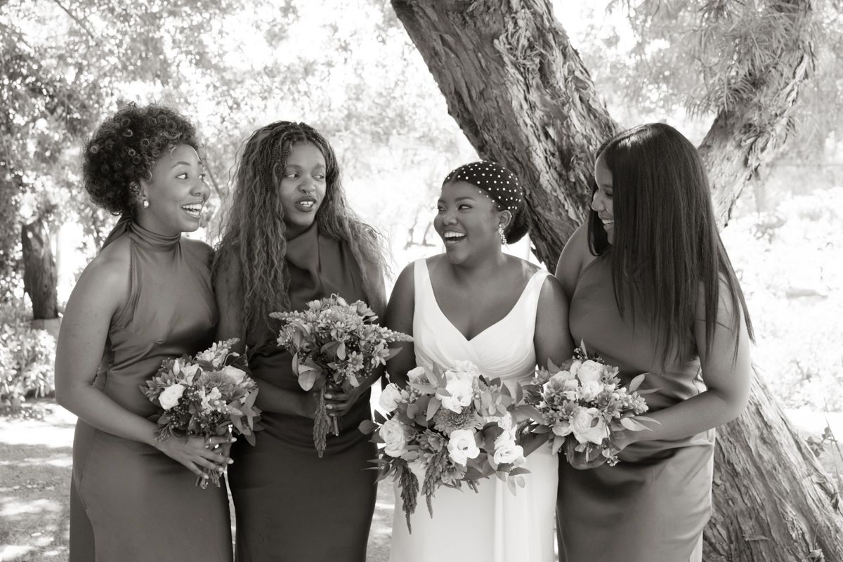 Black and white candid photo of bride and bridesmaids sharing a joyful moment while holding bouquets under a large tree.