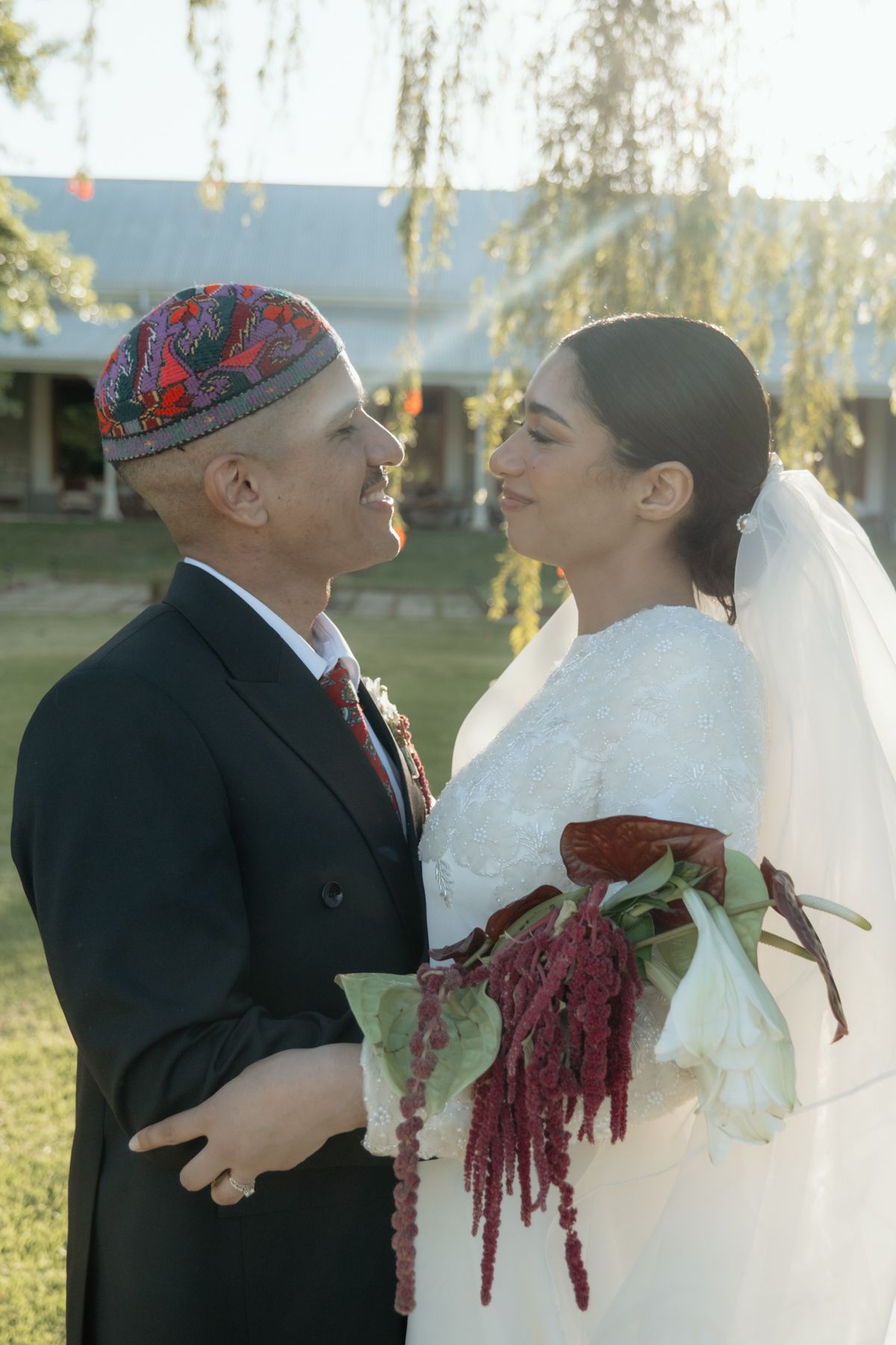 Bride and groom gazing lovingly at each other, groom wearing colorful cap, bride holding red bouquet.