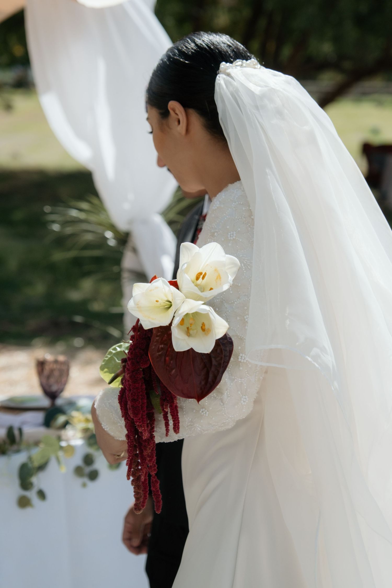 Bride in white lace gown and veil holds bouquet of white tulips and red amaranthus at outdoor wedding.