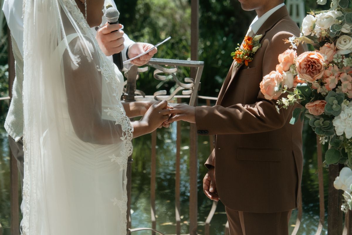Bride and groom exchange vows holding cards under floral arch at outdoor wedding ceremony.