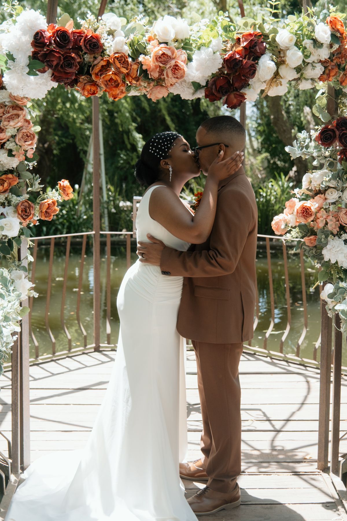 Newlyweds sharing a first kiss under a floral arch adorned with orange, red, and white roses.