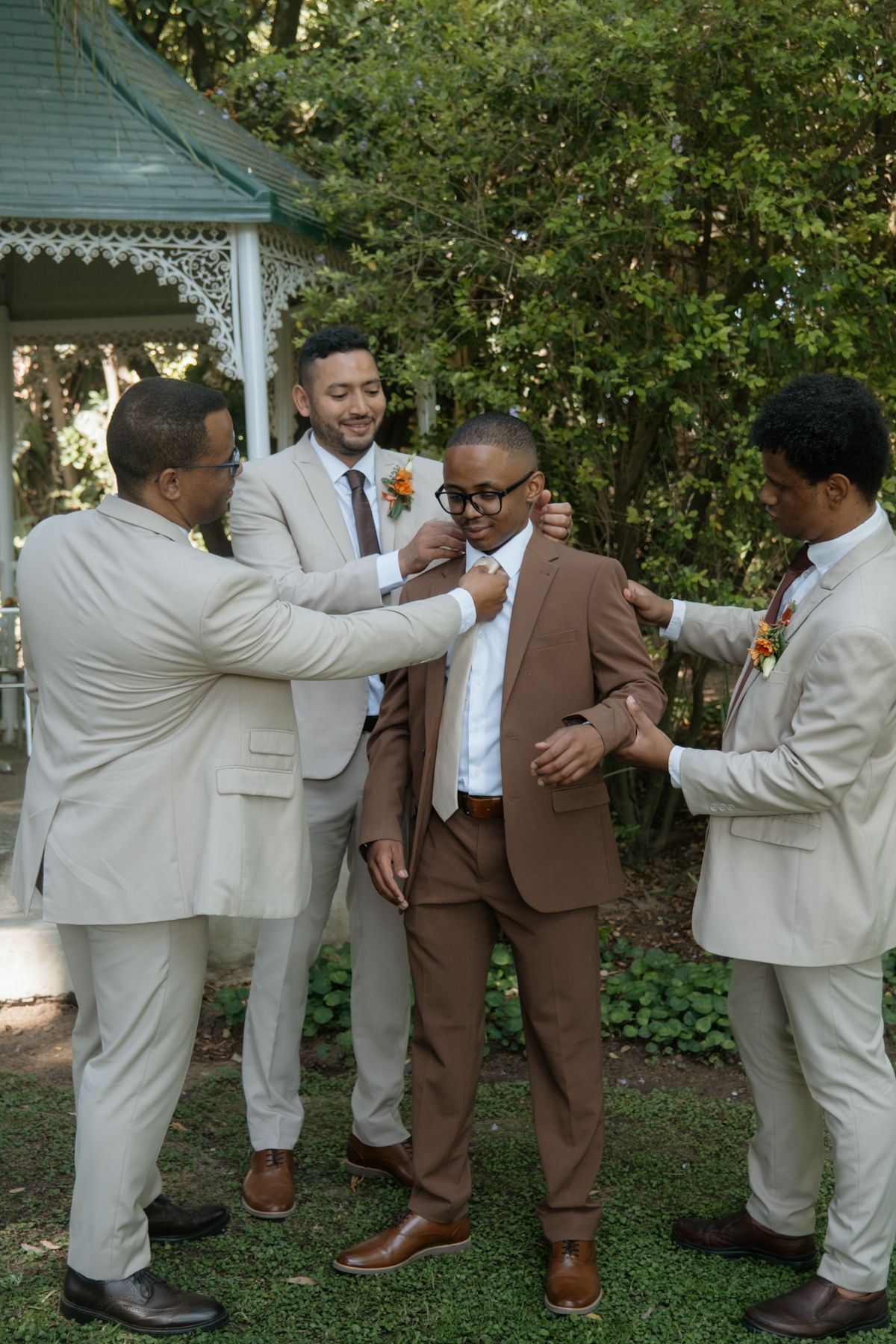 Three groomsmen in tan suits help groom in brown suit with final wedding day preparations outdoors.