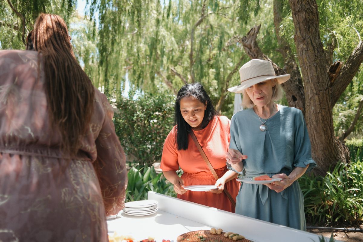 Two women serving food at an outdoor buffet table surrounded by lush green trees on a sunny day.