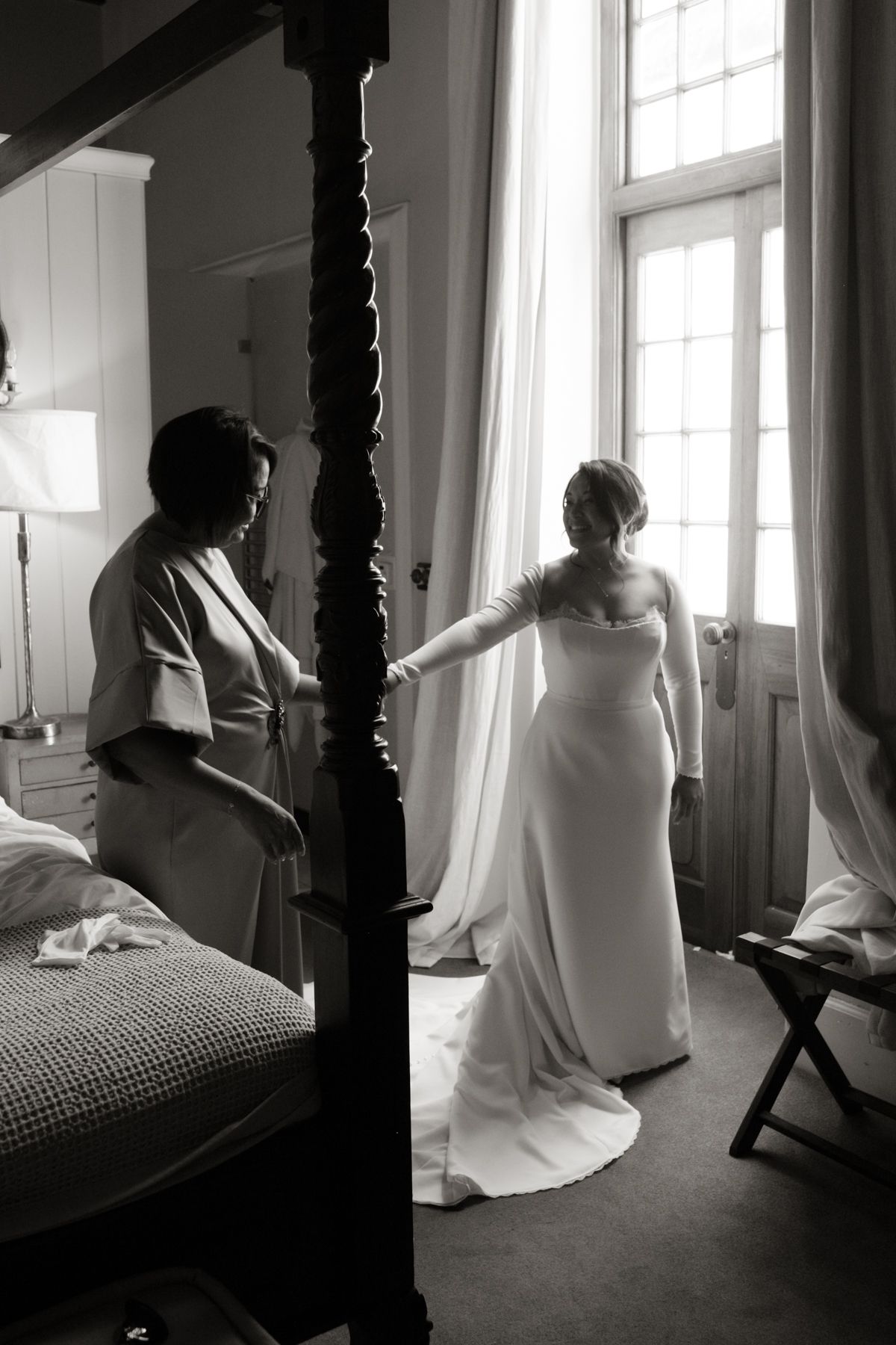 Black and white photo of a bride in a long white gown standing by a window with her mother nearby.