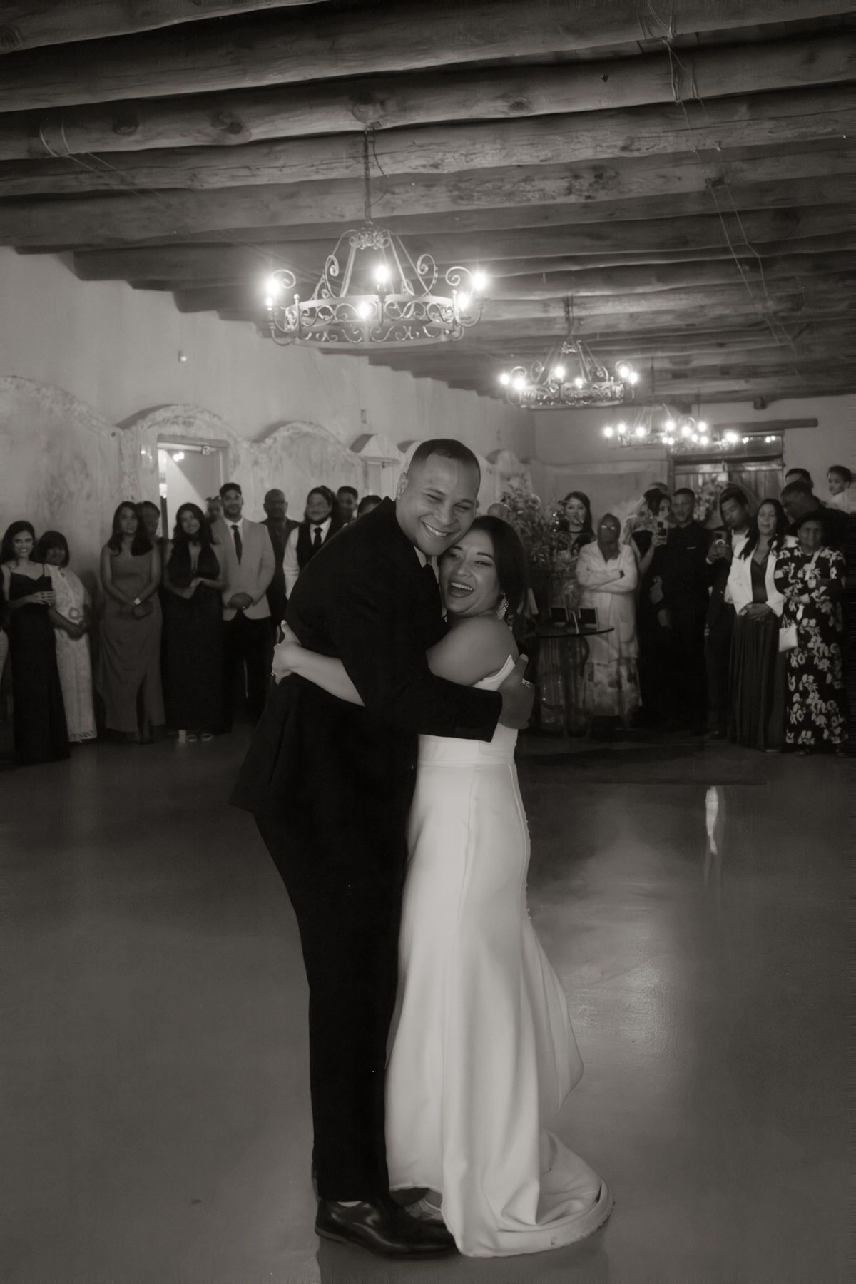 Bride and groom share first dance in black and white photo surrounded by guests in rustic venue.